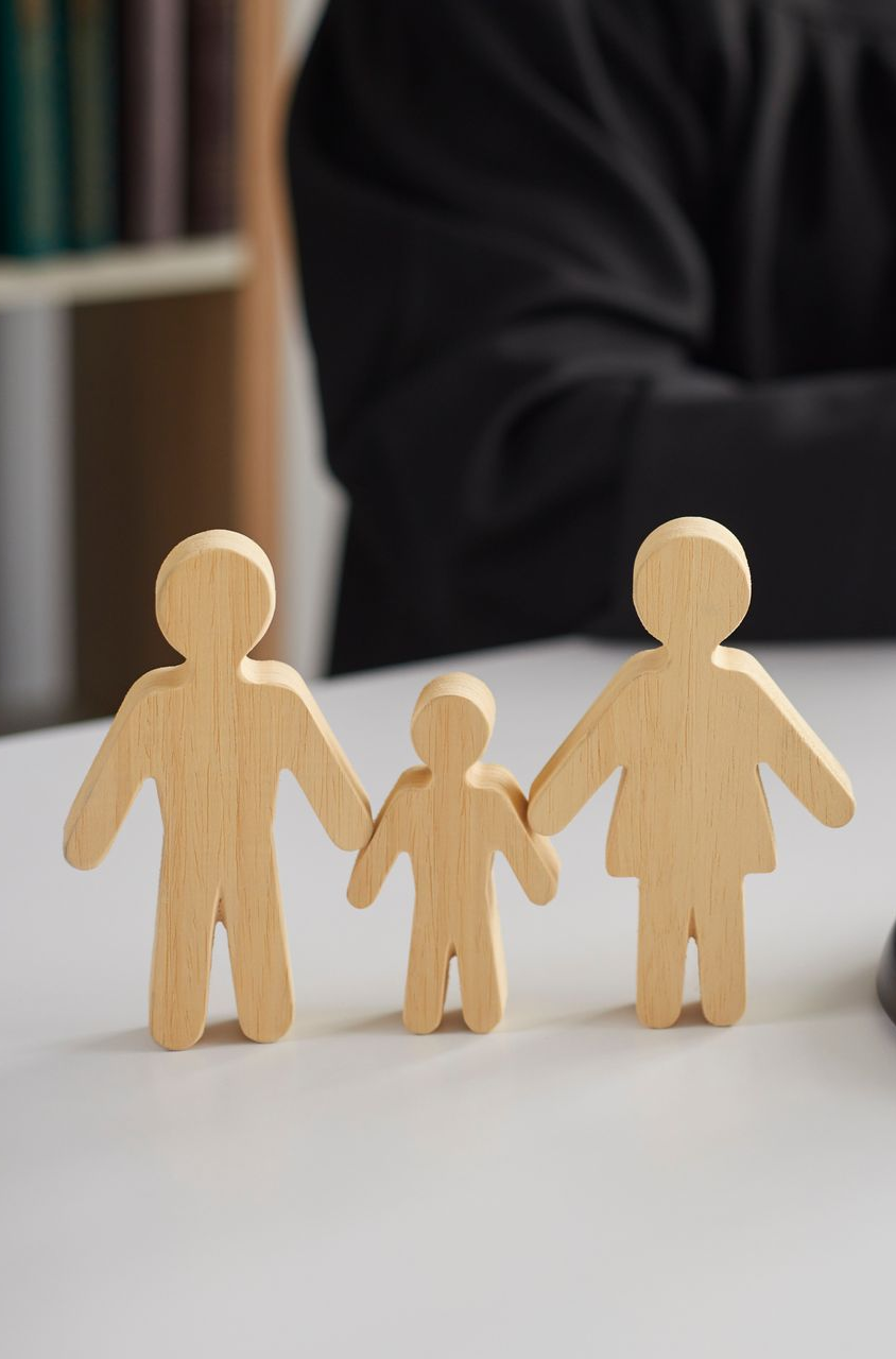 Figurines familiales en bois se tenant la main sur une surface blanche, avec une personne en veste noire en arrière-plan.