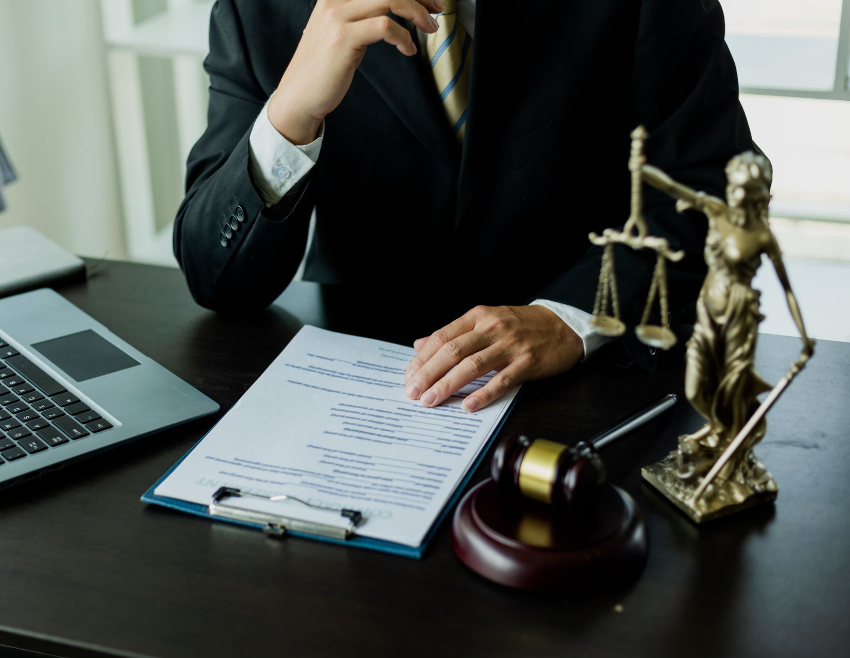 Un avocat en costume, assis à son bureau, examine des documents, avec une balance de la justice, un marteau et un ordinateur portable.