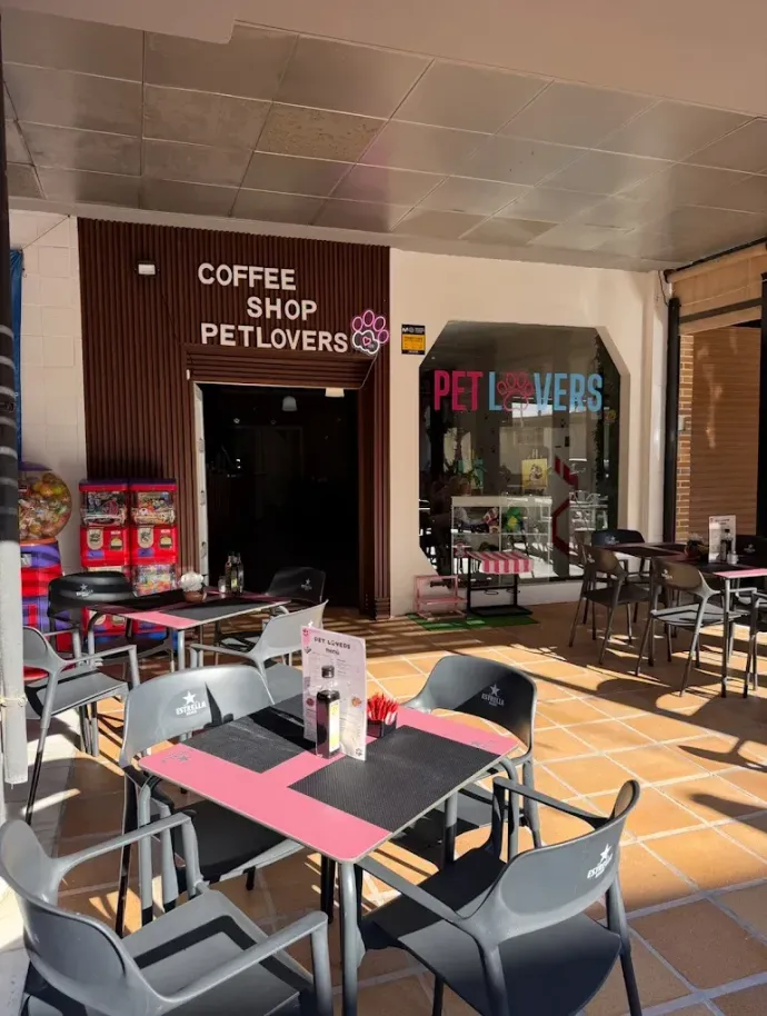Terraza exterior de una cafetería para amantes de las mascotas con mesas, sillas y letreros, bañada por la luz del sol.
