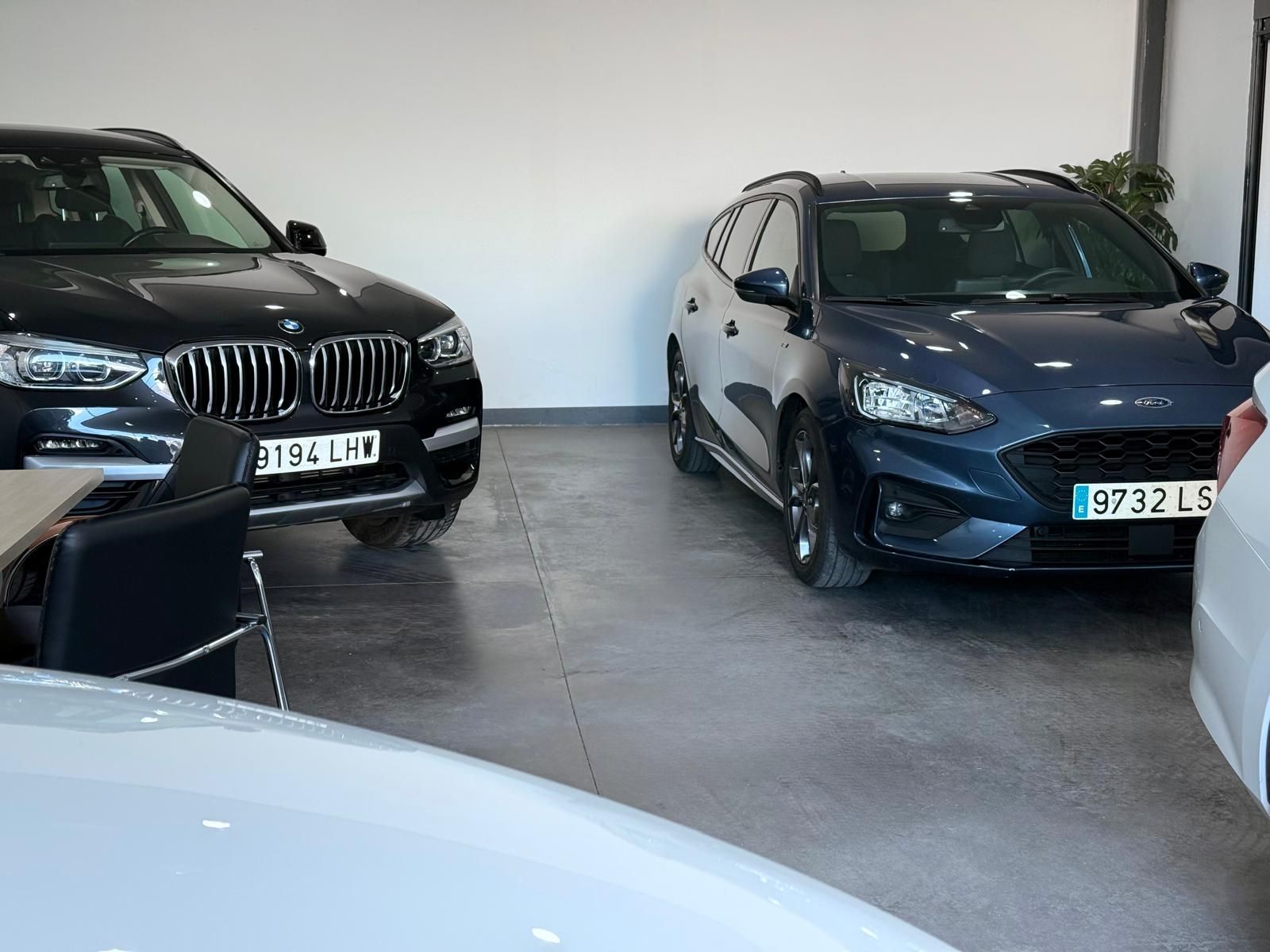 Autos dentro de un showroom: un SUV BMW negro y un Ford Focus azul, piso de concreto, paredes blancas.