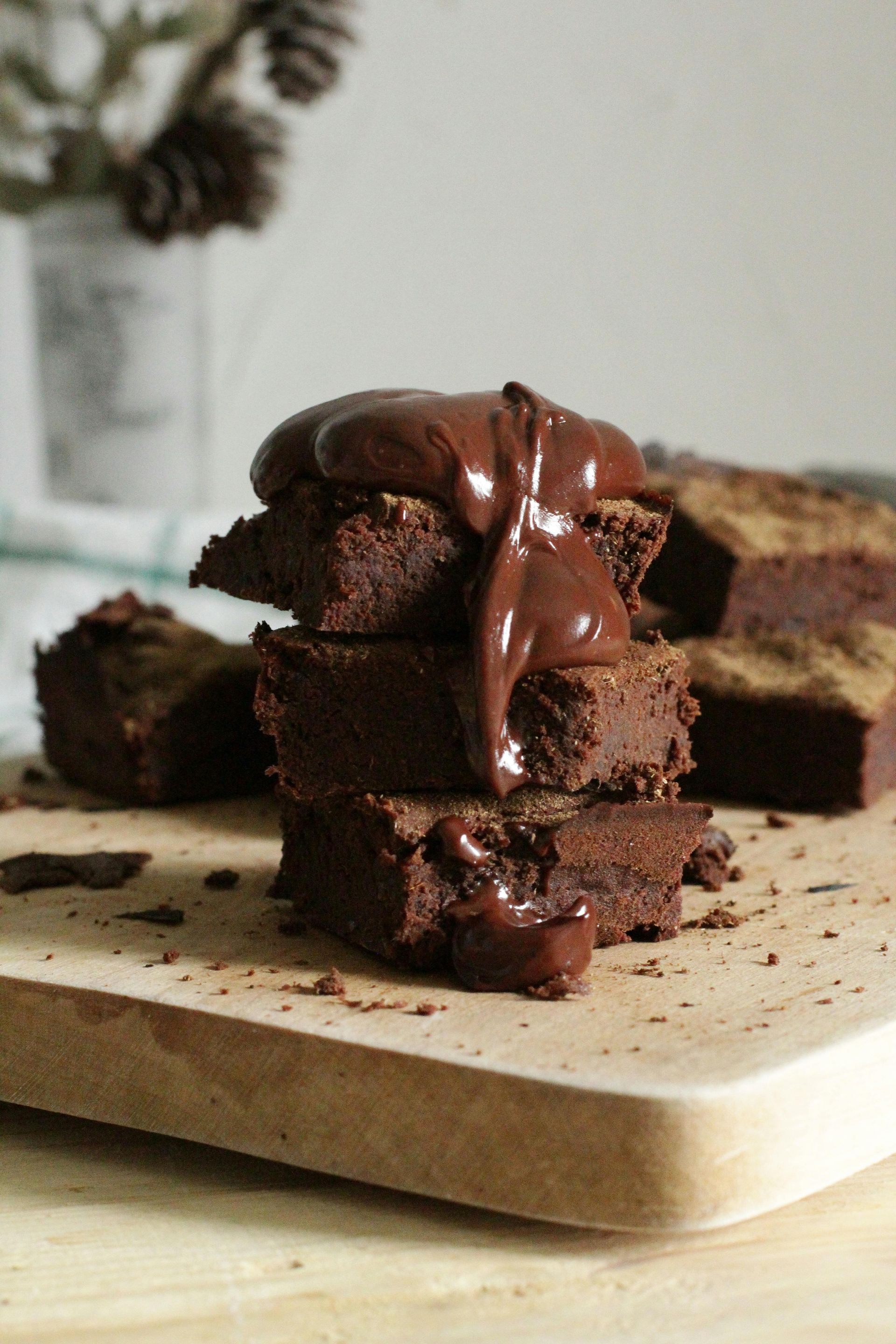 Pila de brownies con salsa de chocolate sobre una tabla de madera; otros brownies en el fondo, piñas.