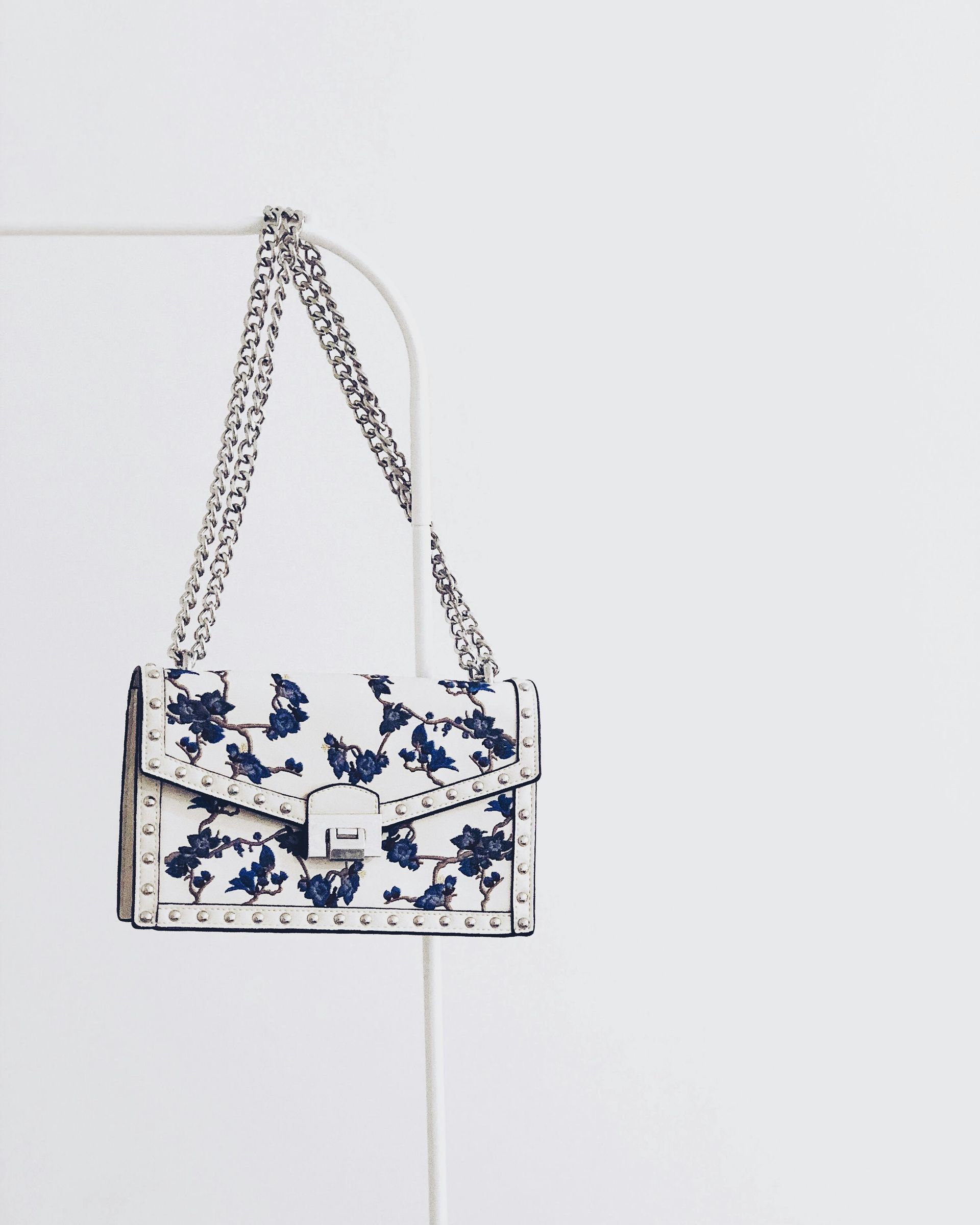 Bolso blanco con estampado floral azul, correa de cadena plateada, colgado sobre un fondo blanco.