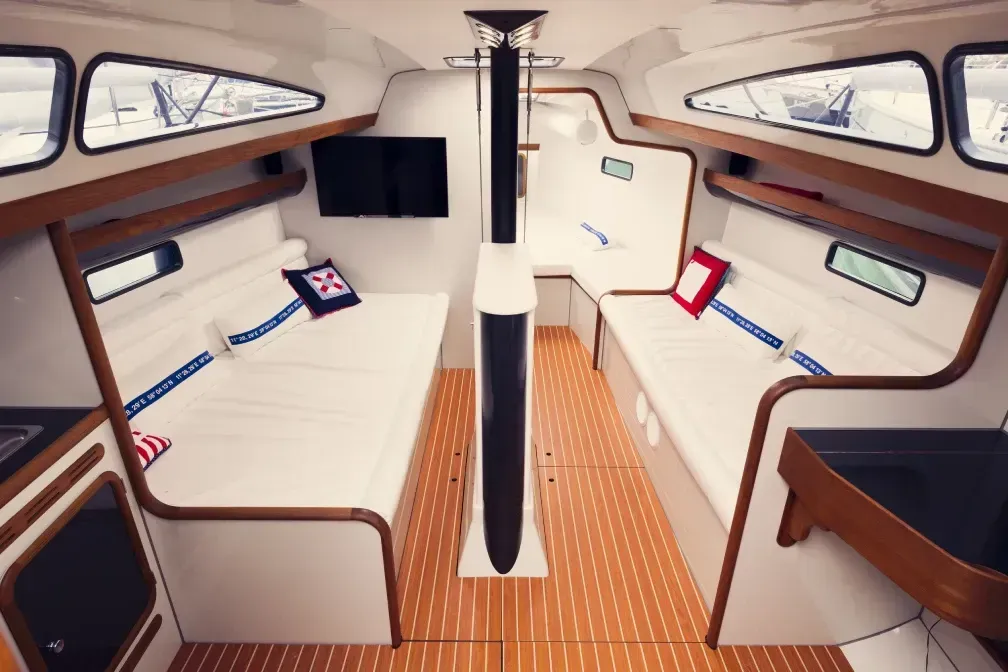 Interior de la cabina de un velero, con asientos blancos, pisos de madera y un mástil central.