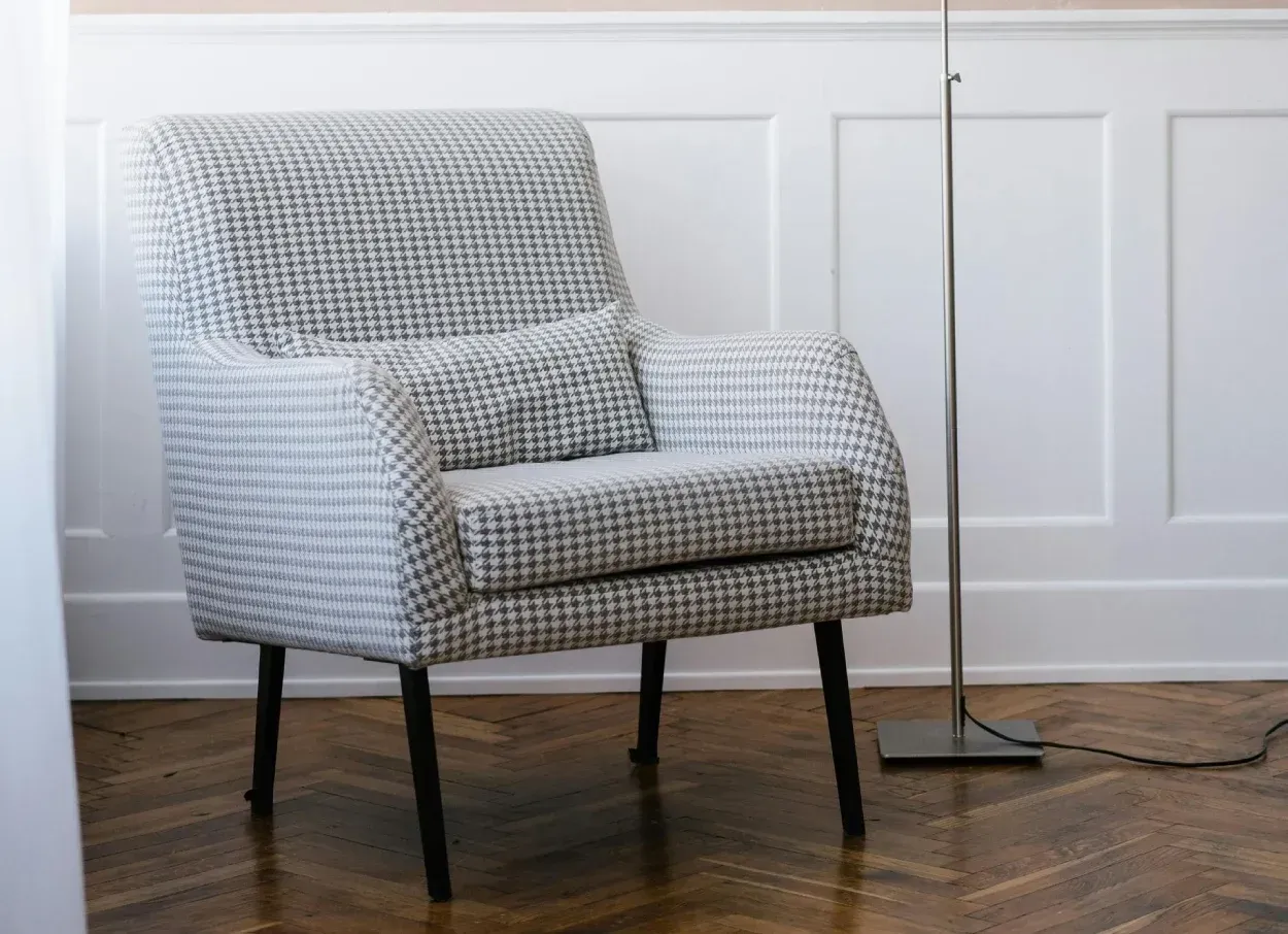 Sillón con tejido estampado y patas negras, con almohada, contra pared de paneles blancos, lámpara de pie.