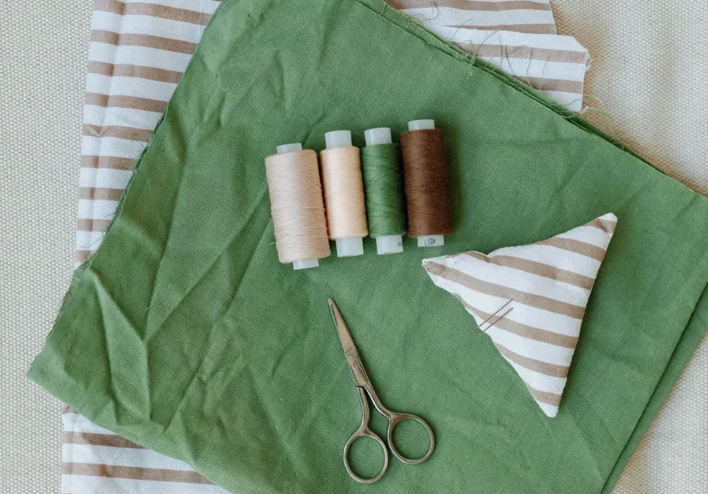 Tela verde, carretes de hilo, tijeras y una figura de tela rayada, dispuestas para coser.