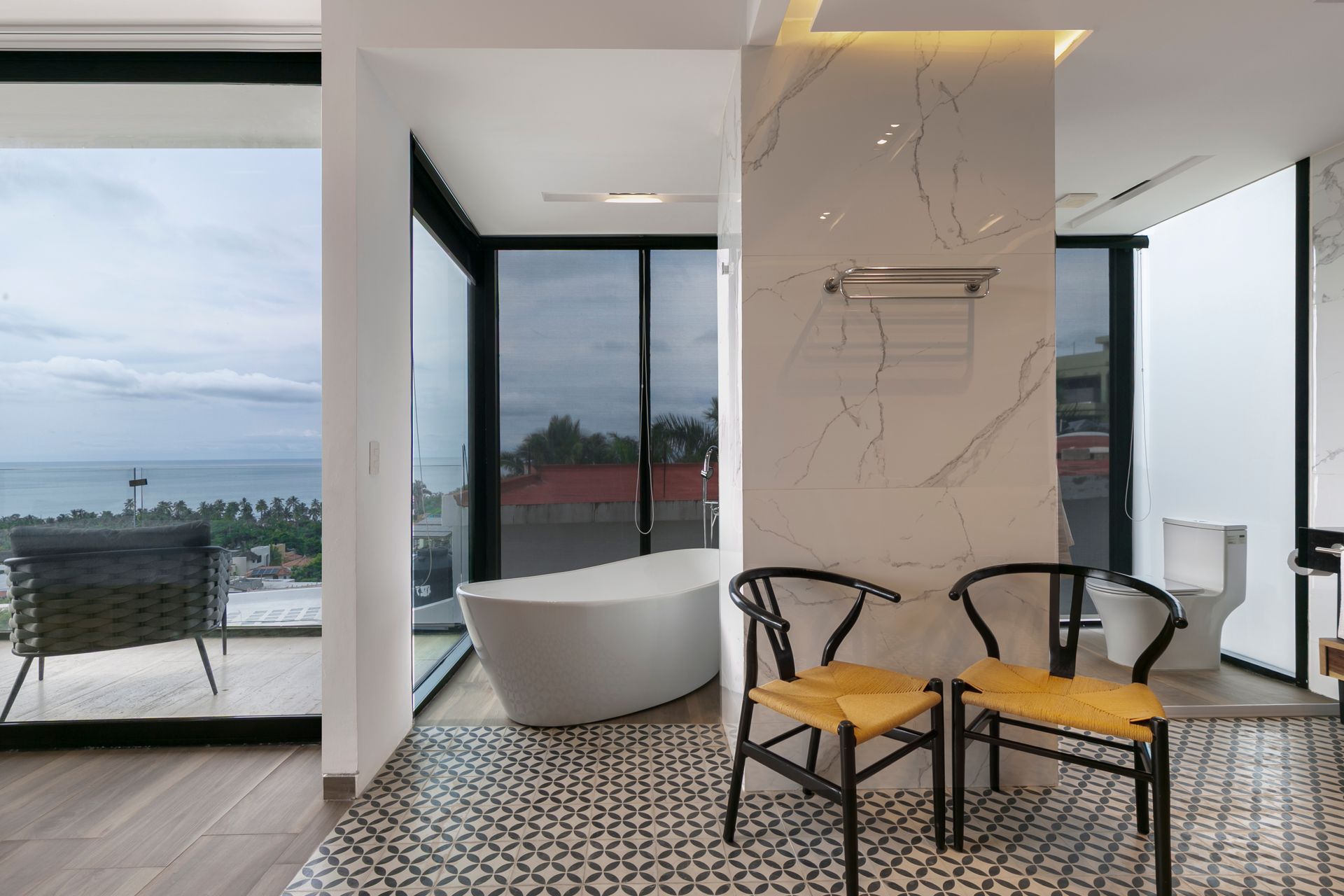 Modern bathroom with a white tub, ocean view, patterned floor, and two chairs.