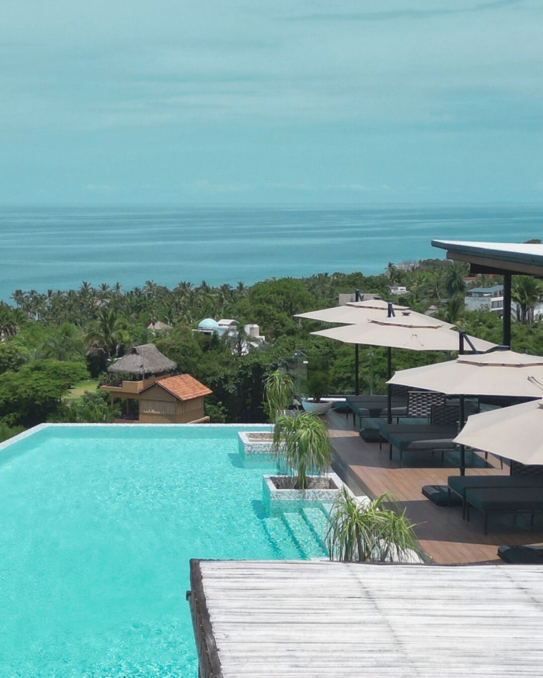 Poolside view overlooking ocean, turquoise water, wooden deck, lounge chairs with umbrellas.