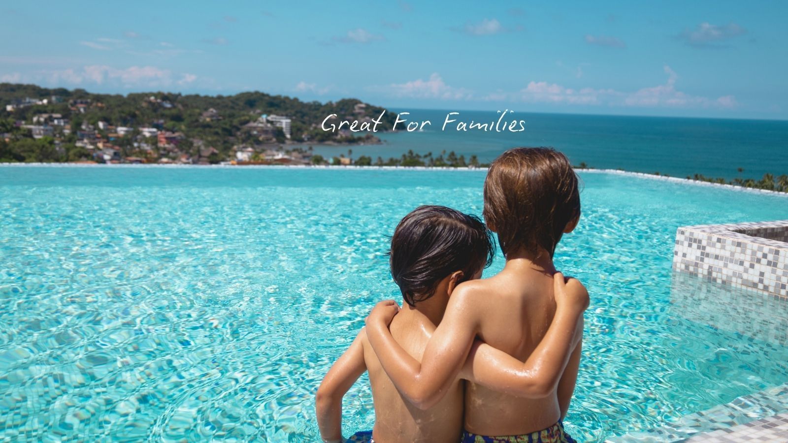 Two young boys in swimsuits embrace, looking out at an infinity pool and ocean.