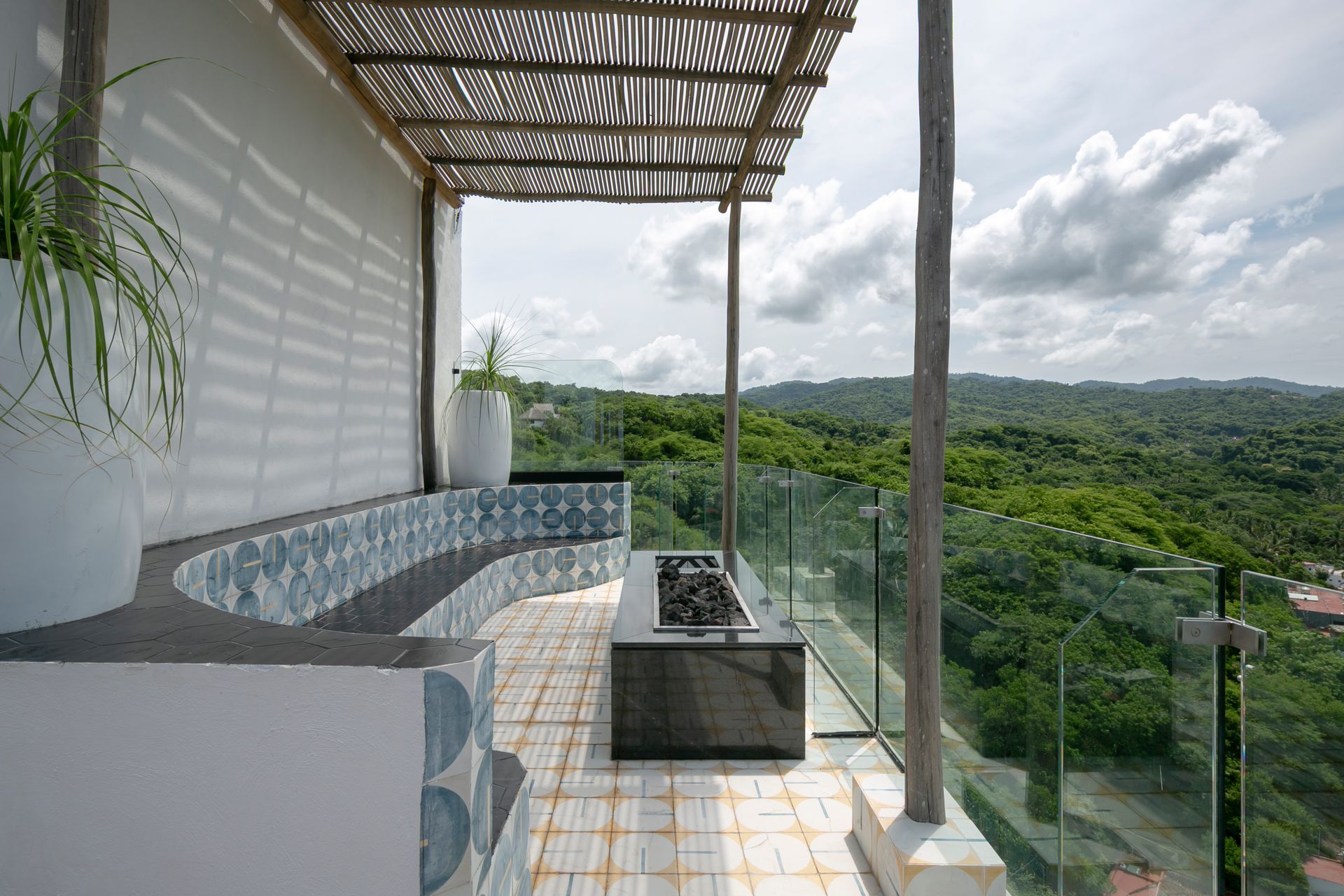 Outdoor patio with glass railing, bench, and forest view, shaded by a wooden pergola.