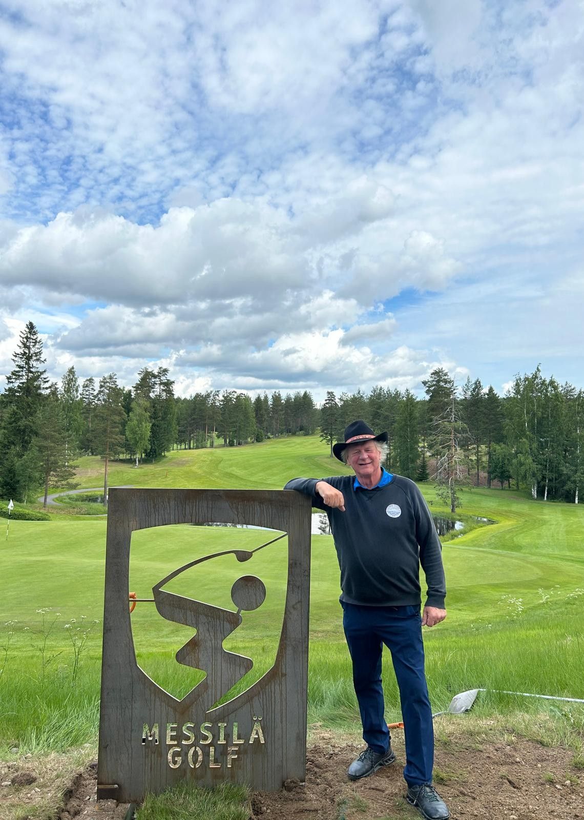 Mies seisoo Messilan golfkentän kyltin vieressä vihreällä väylällä pilvisen taivaan alla