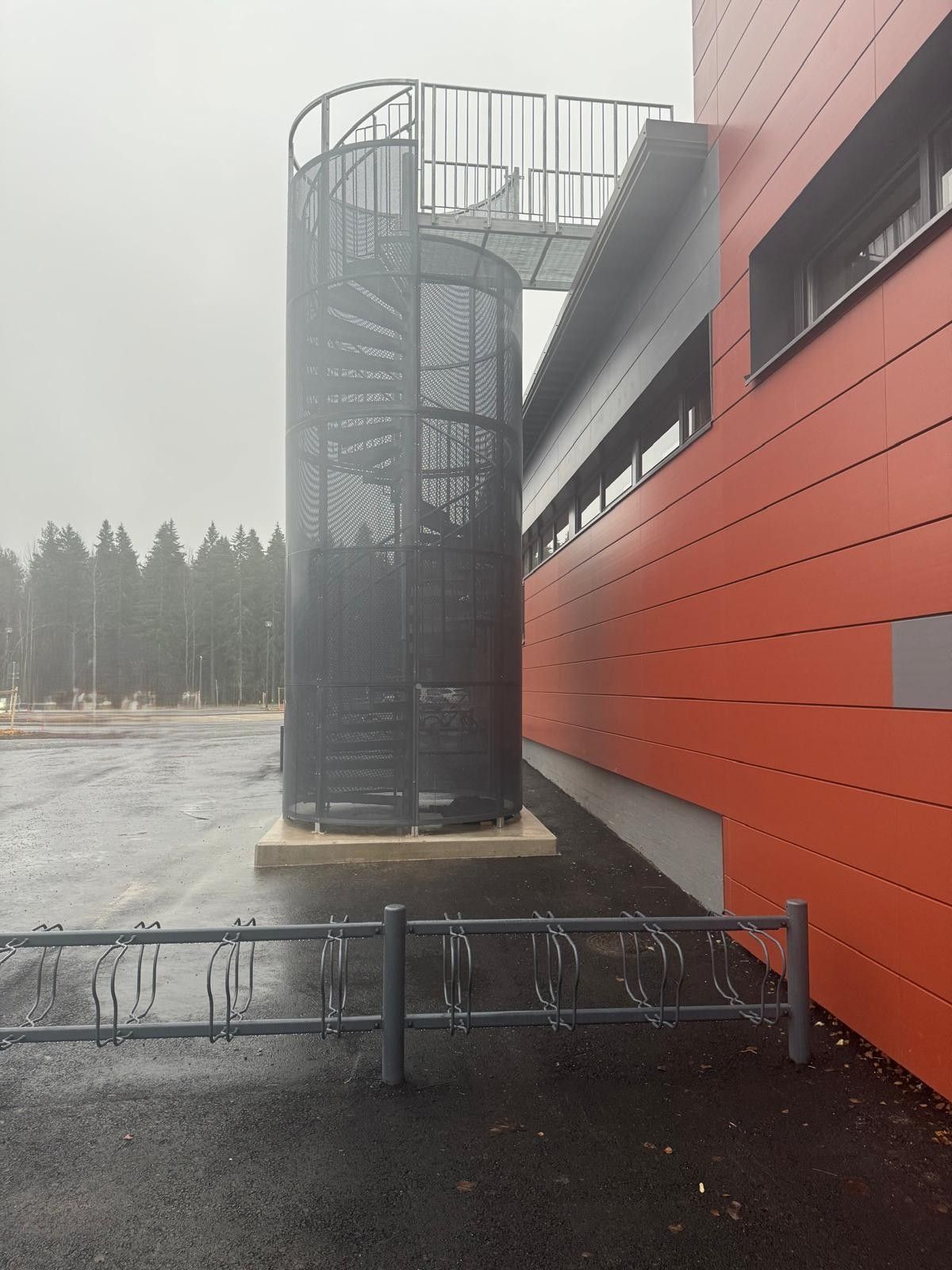 Spiraalimainen paloportaat punaisen rakennuksen vieressä sateisella, tyhjällä tontilla, jonka edessä on kaatunut metalliaita