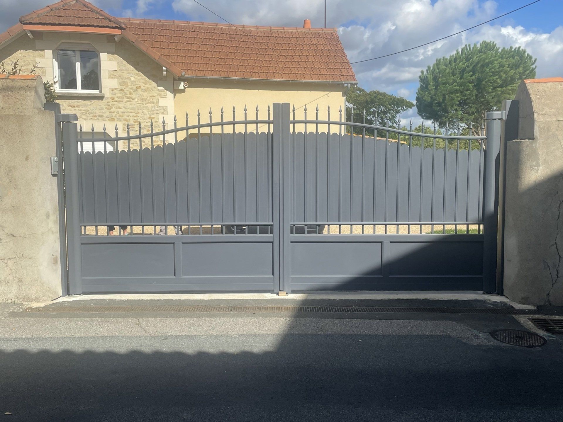 Portail en métal gris devant une maison beige au toit de tuiles rouges.