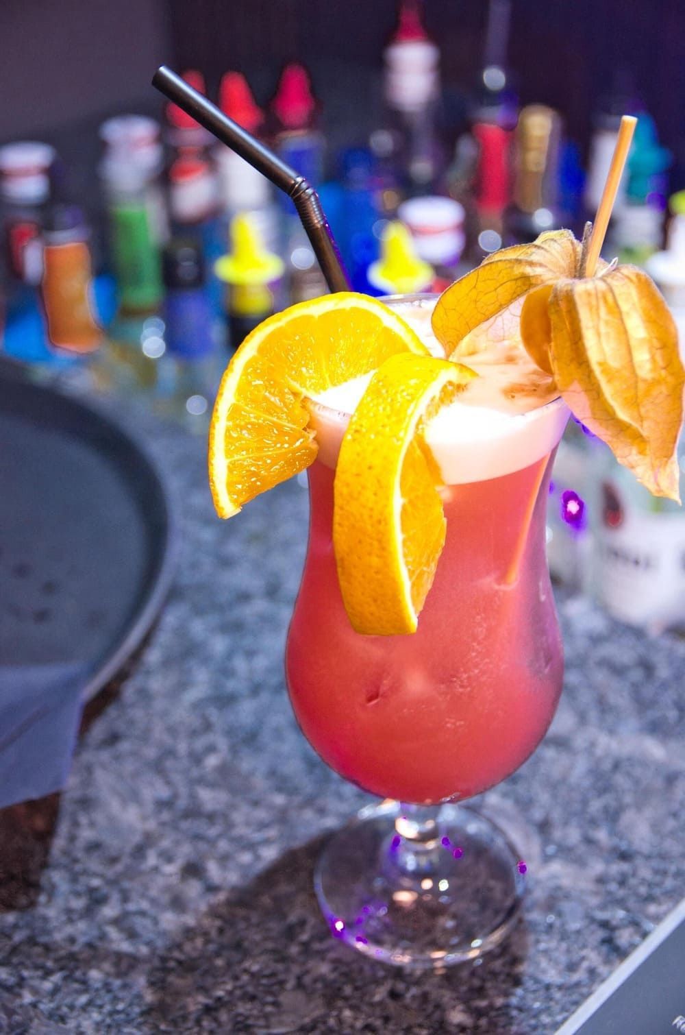 Ein roter Cocktail mit Orangenscheiben und einem Strohhalm auf einer Bar | By Mikey