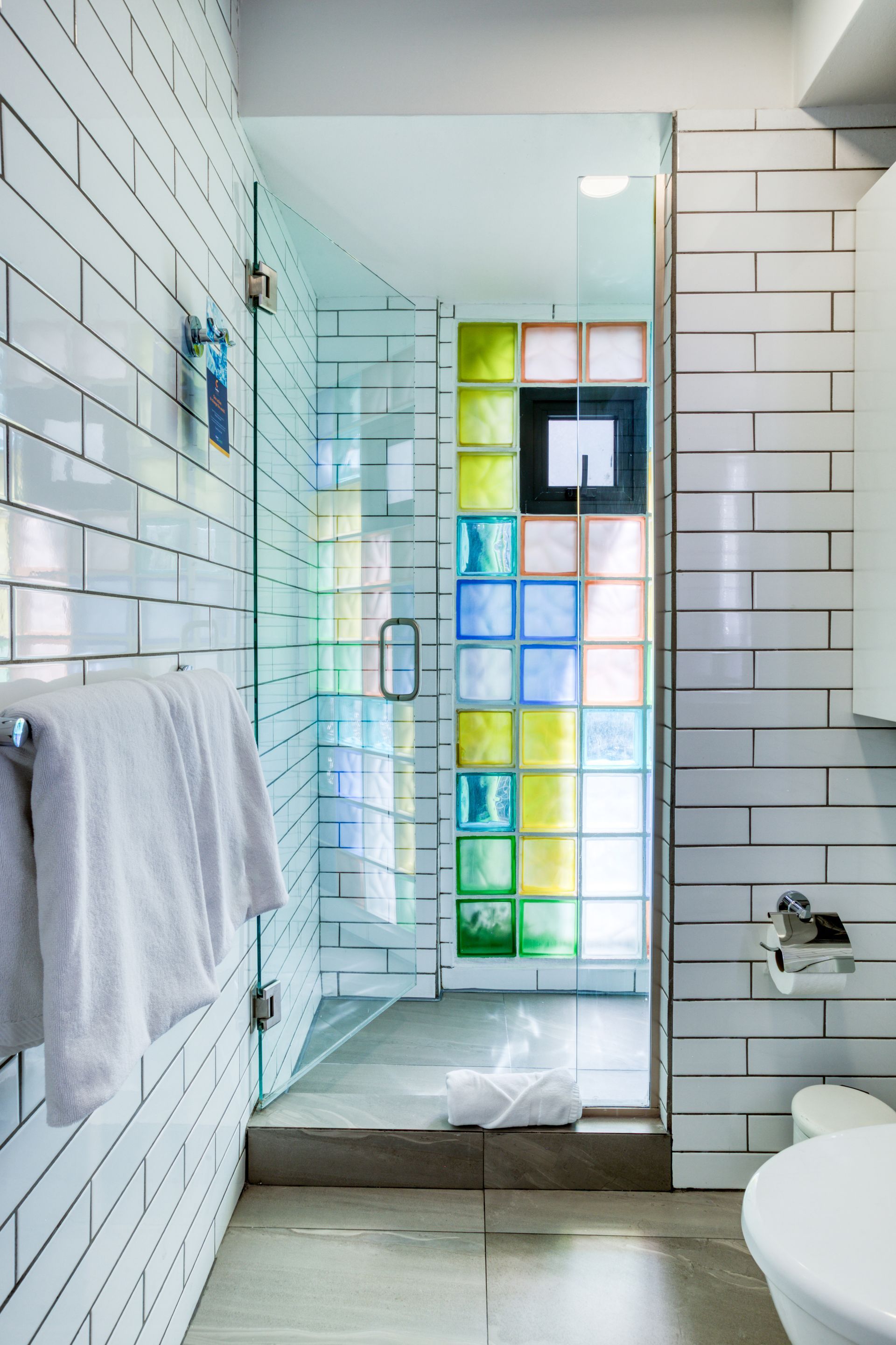 Modern bathroom with white subway tiles, a multicolored glass block shower wall, white vanity, and open shelving.