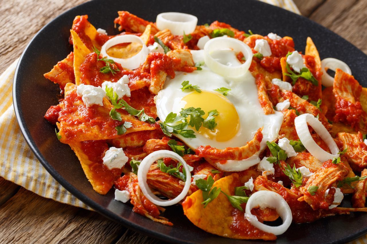 Chilaquiles with fried egg, red sauce, cheese, onions, and cilantro on a black plate.