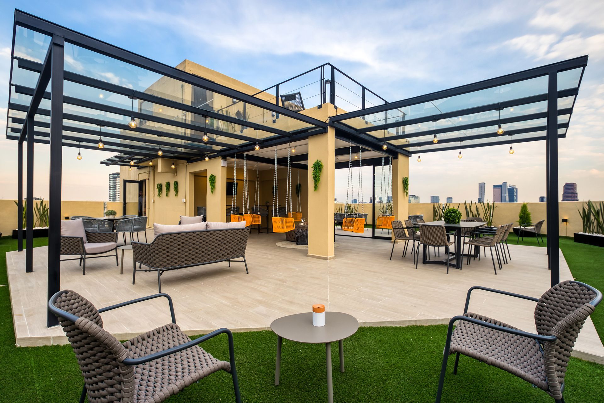 Rooftop patio with seating, glass canopy, and string lights, overlooking a city skyline.