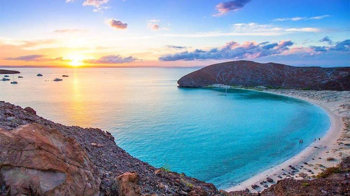 Sunset over a turquoise bay, beach, boats, and dark cliffs.