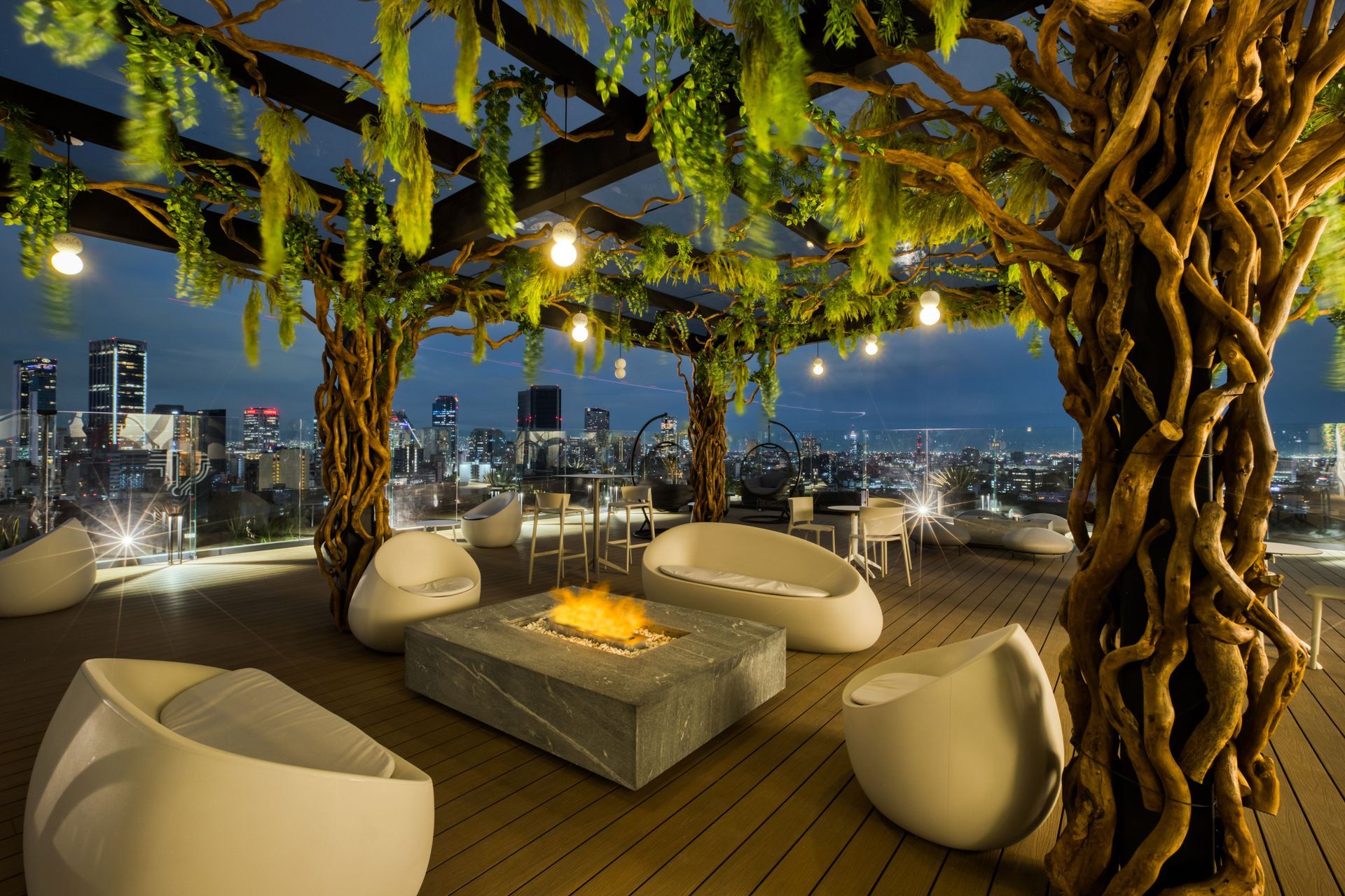 Rooftop patio with city view, lounge chairs around fire pit, tree-like pillars, and string lights.