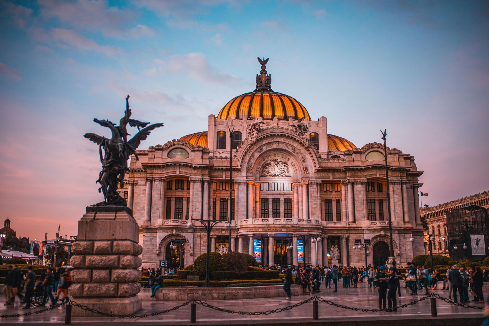 Palacio de Bellas Artes in Mexico City at sunset; gold dome and ornate architecture; statue in foreground, crowd.
