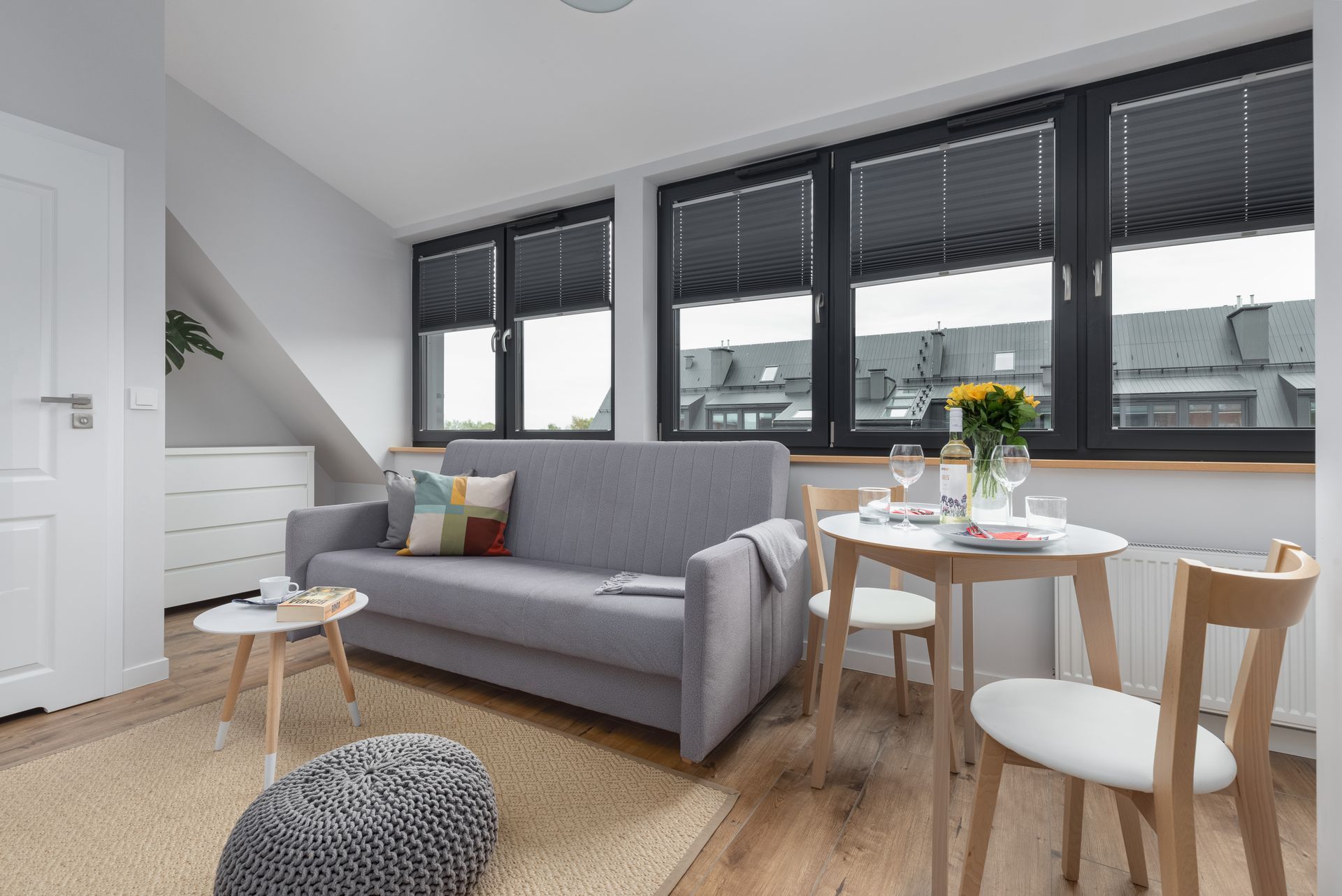 Mały, jasny salon z szarą sofą, okrągłym stołem dla dwóch osób i dużymi oknami z zasłoniętymi roletami.