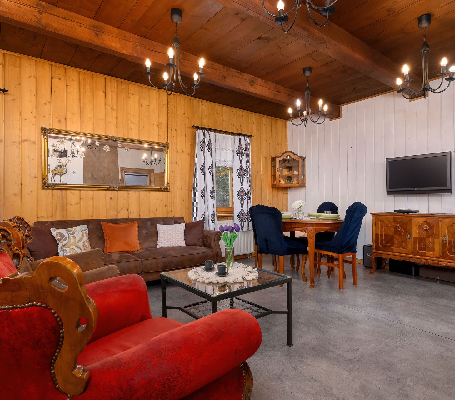 Salon z boazerią, sofą, czerwonym krzesłem, stołem jadalnym i żyrandolami.