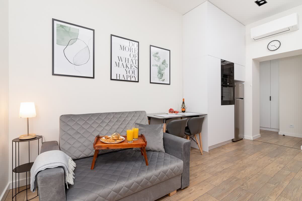 Przytulny salon z szarą kanapą, małym stolikiem, dziełami sztuki i aneksem kuchennym.
