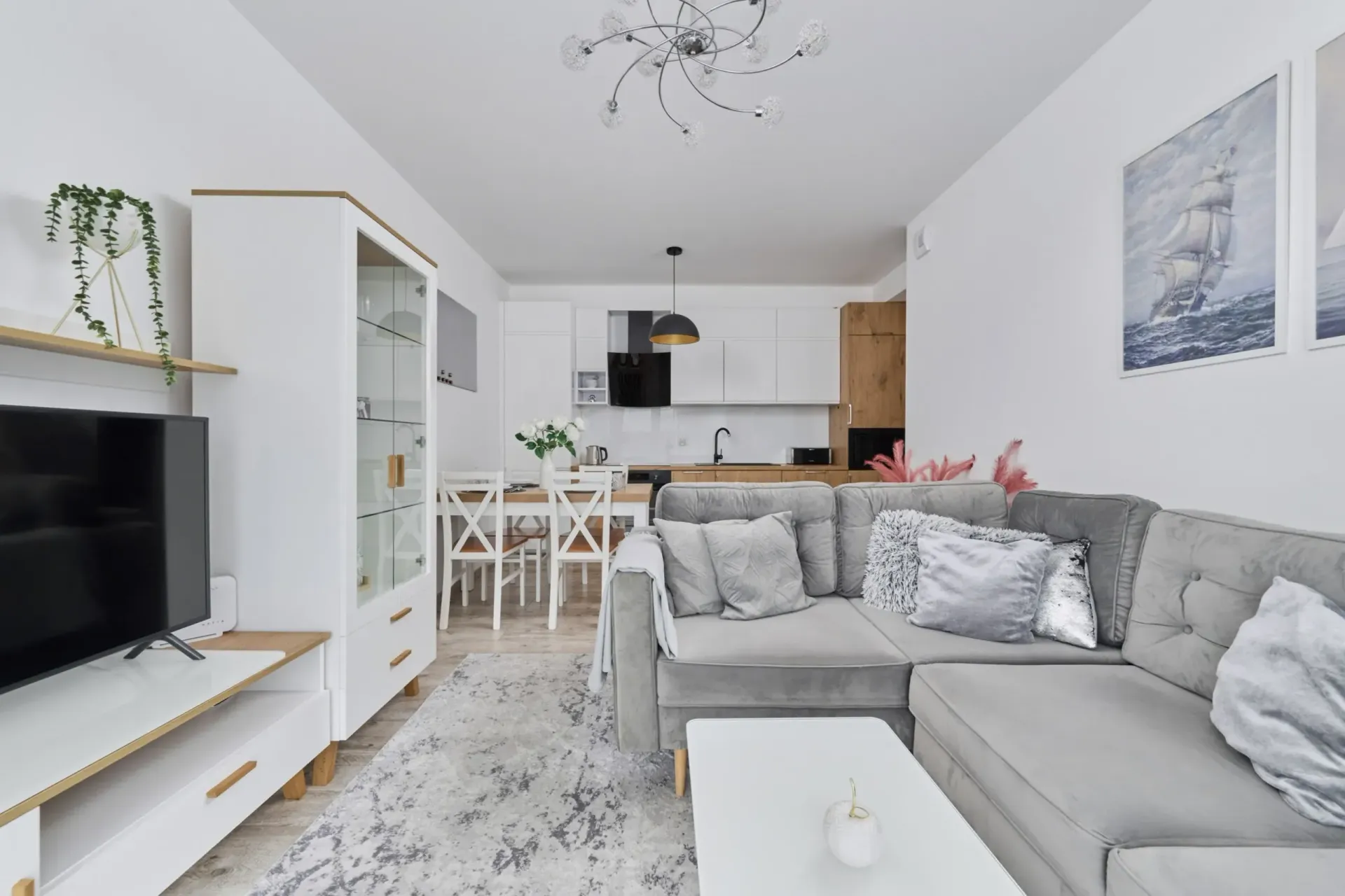 Salon z szarą narożną sofą, białymi szafkami i otwartą kuchnią.