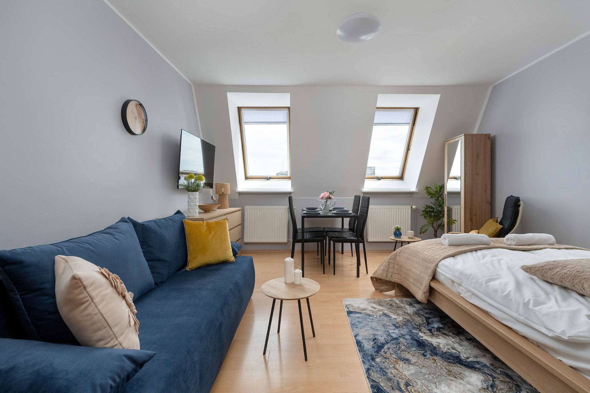 Apartament typu studio z niebieską sofą, łóżkiem, stołem jadalnym i dwoma oknami dachowymi.