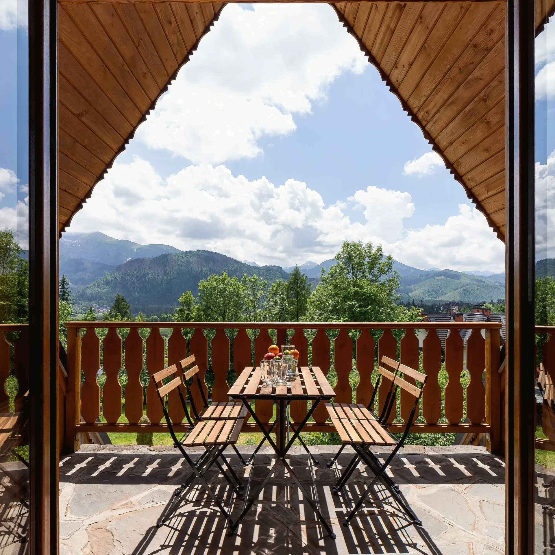 Drewniany balkon ze stołem i krzesłami, z widokiem na góry i zachmurzone niebo.
