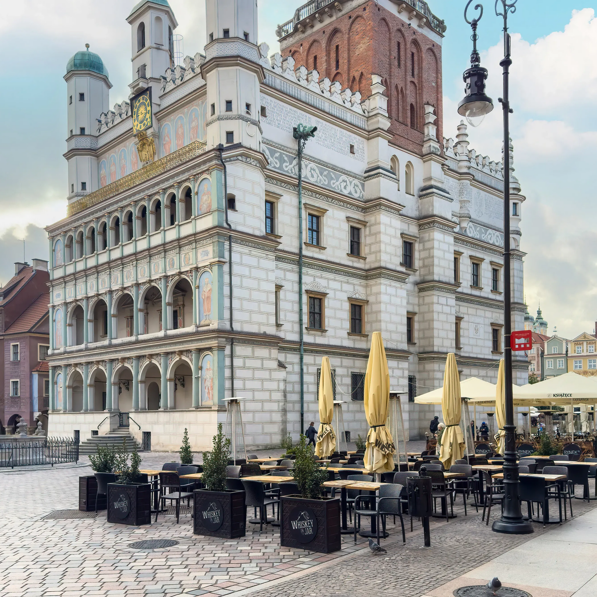 Budynek z miejscami do siedzenia na zewnątrz, na placu. Biały i beżowy kamień z czerwoną wieżą, parasolami i ludźmi.