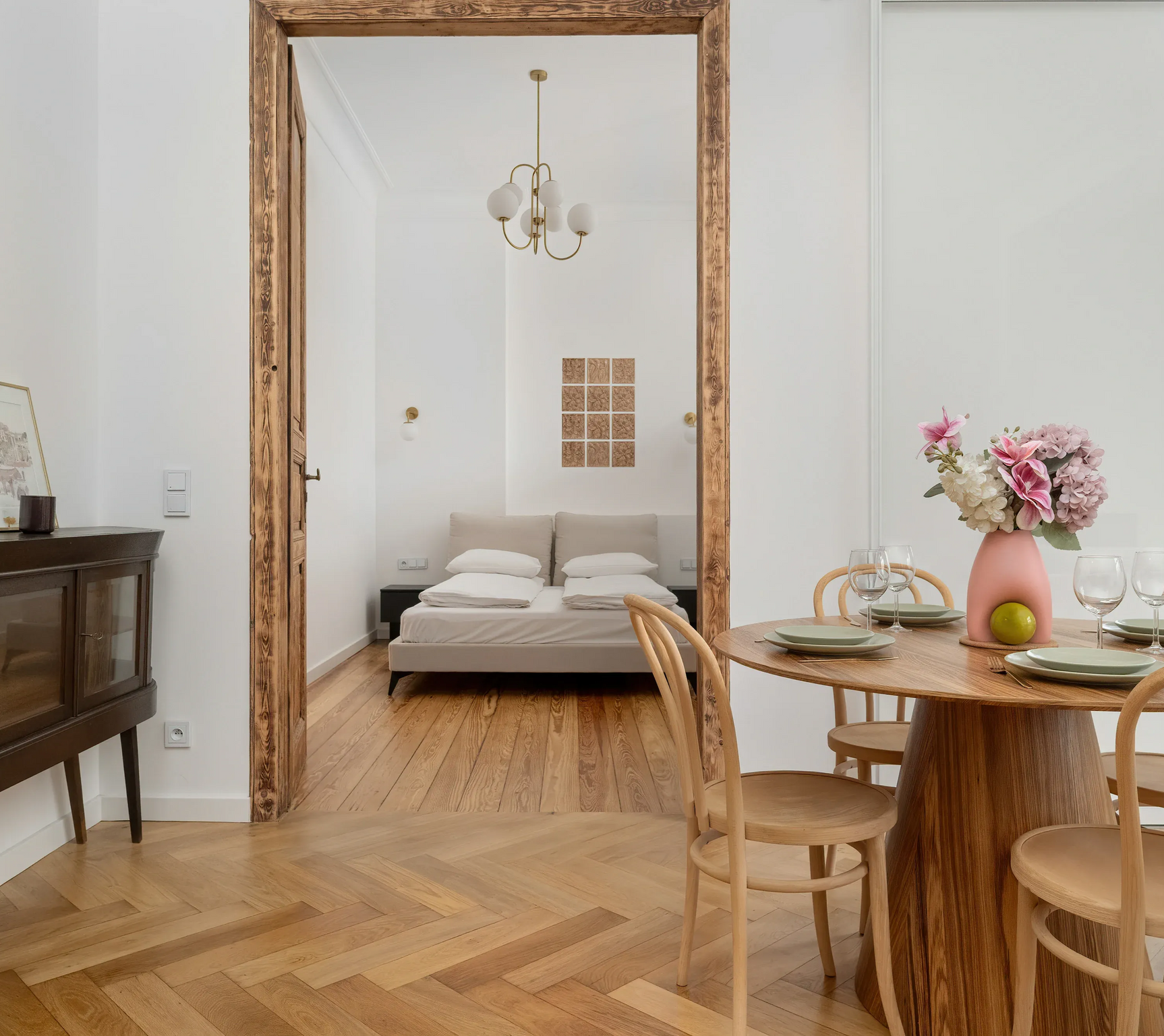 Interior with wooden floors, dining table, and bedroom in the background. Pink vase with flowers, antique cabinet.