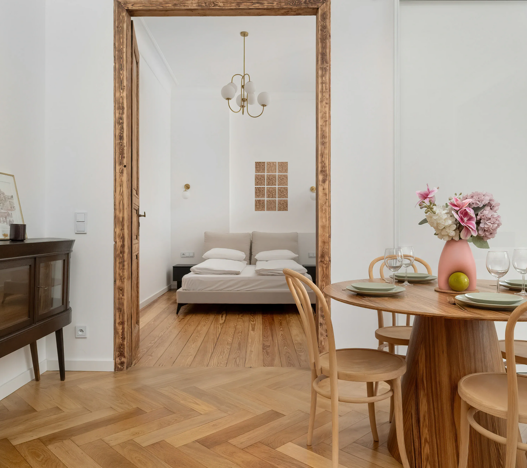 Widok przez drewniane drzwi do sypialni z łóżkiem i lampą; na pierwszym planie znajduje się jadalnia.