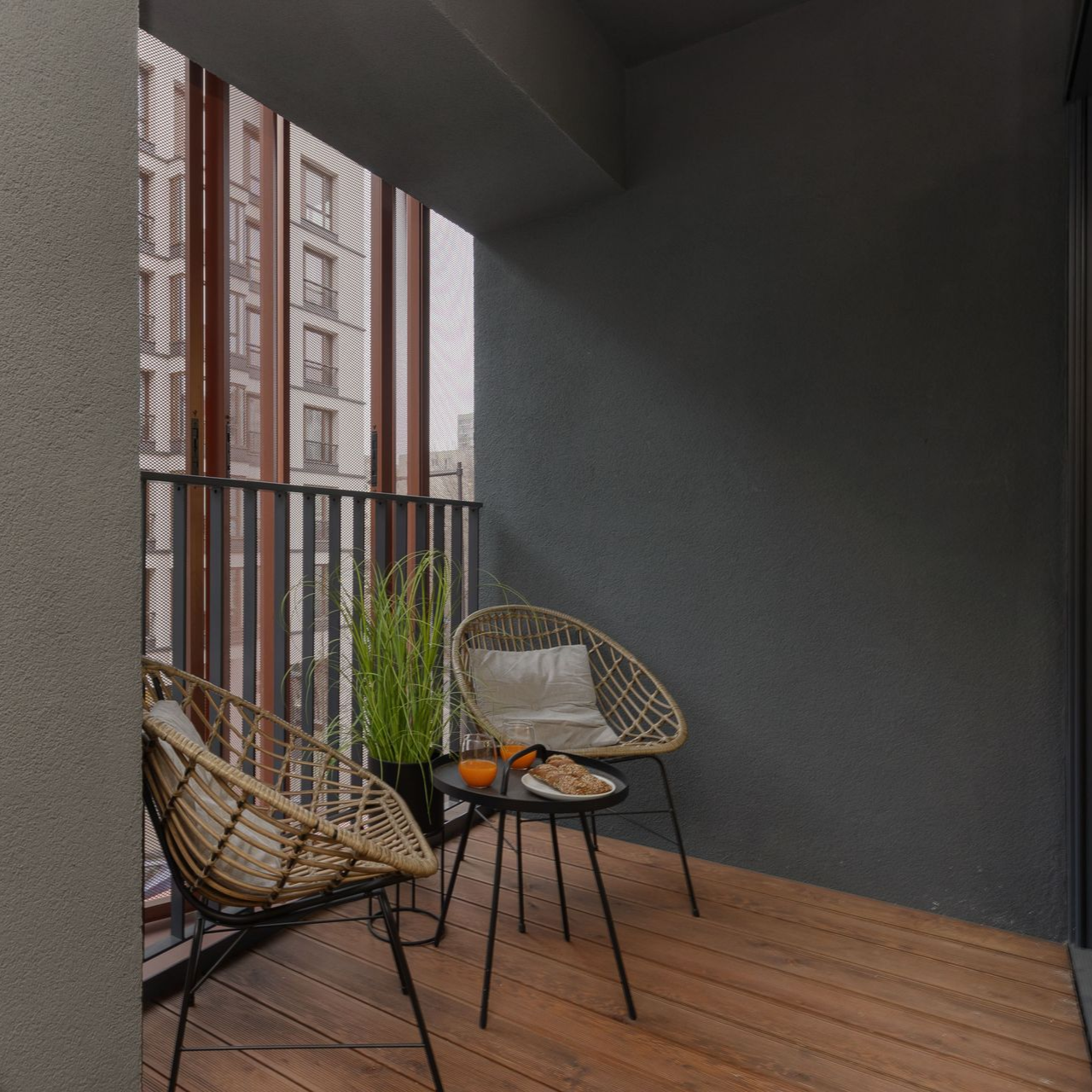 Mały balkon z dwoma wiklinowymi krzesłami, małym stolikiem z jedzeniem/napojami i widokiem na miejskie budynki.
