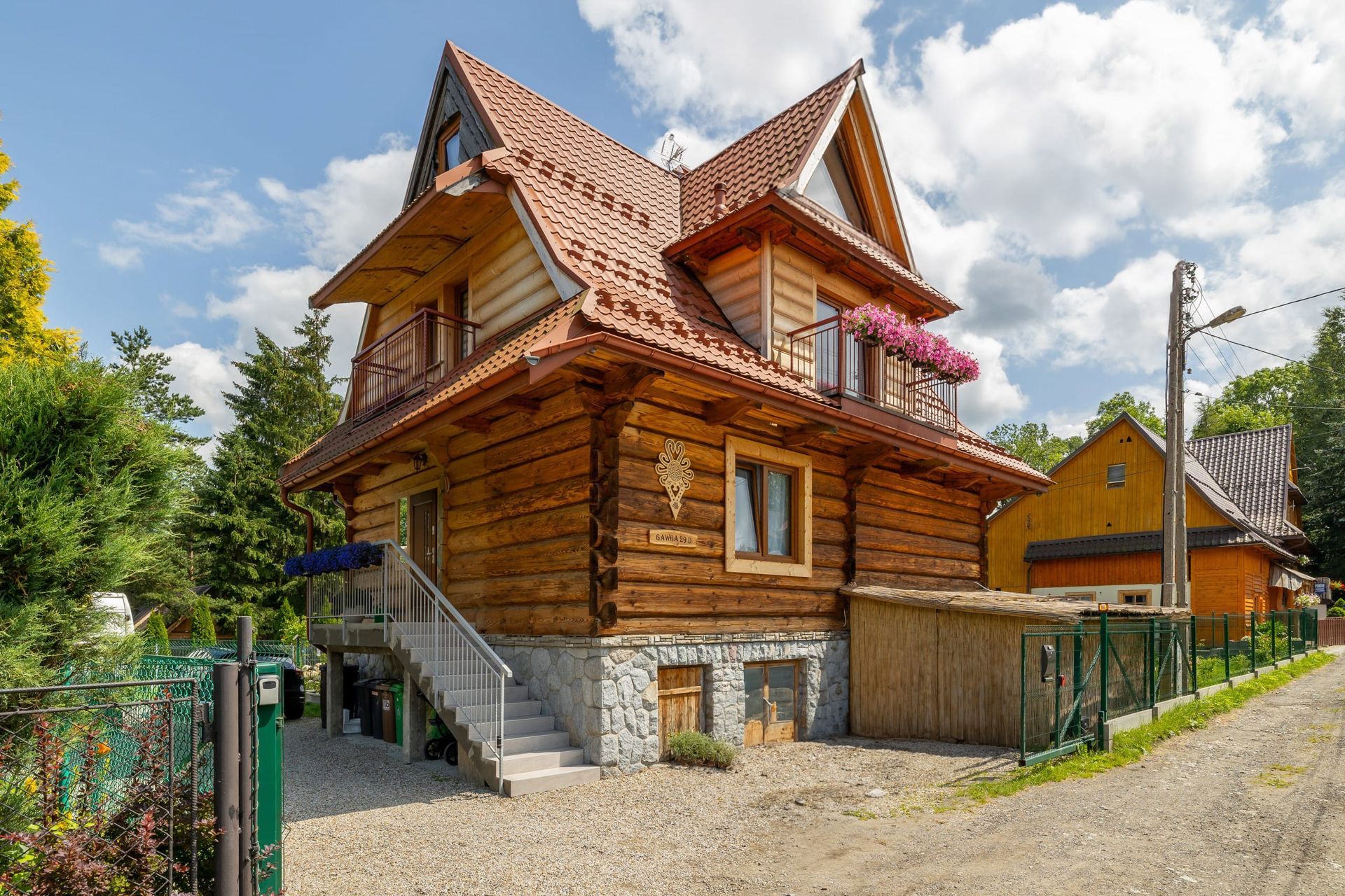Drewniany domek z brązowymi balami i czerwoną dachówką, kamiennymi fundamentami i szarymi schodami. Dom posiada balkony z kwiatami.