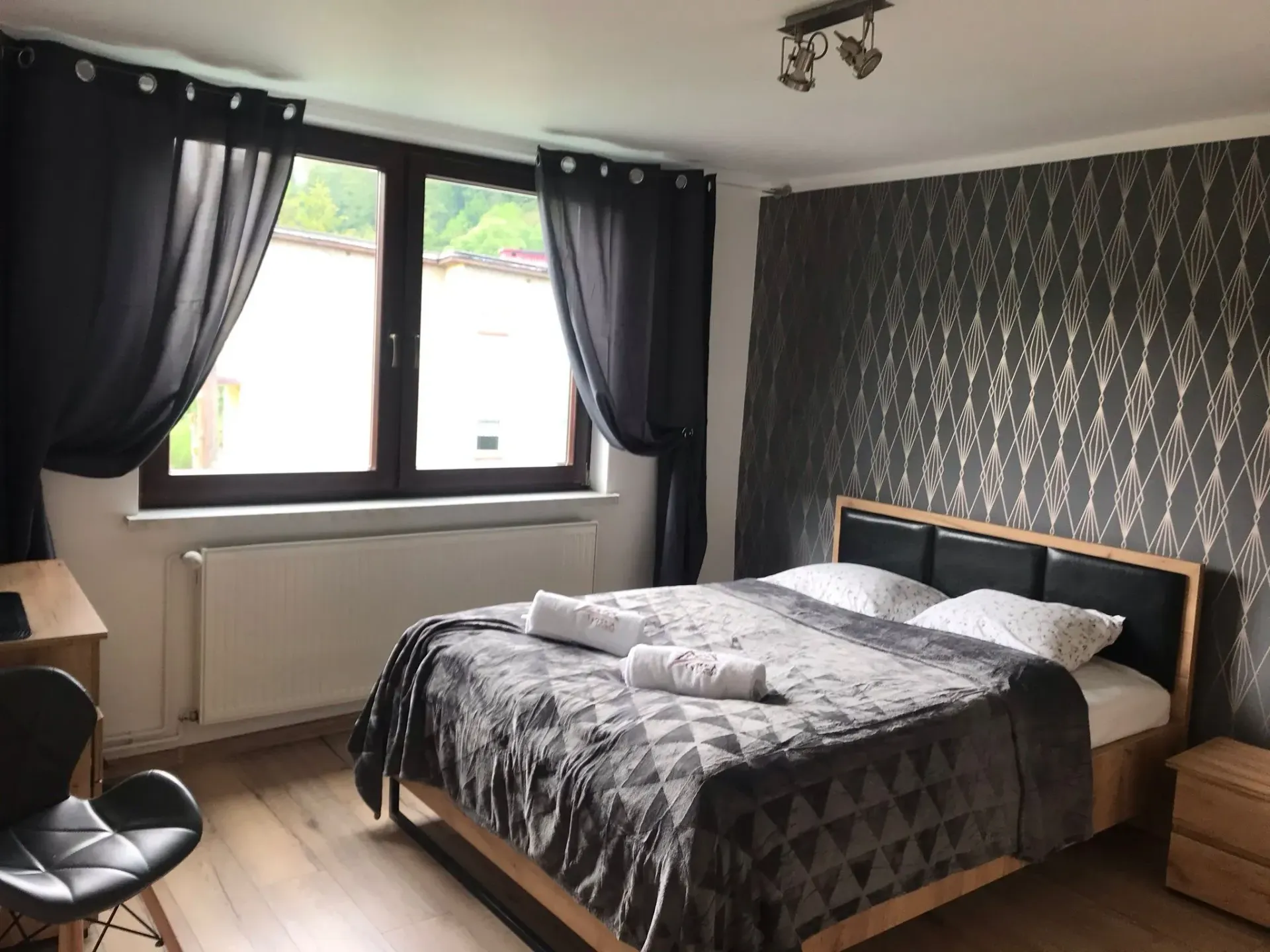 Sypialnia z łóżkiem, oknem z ciemnymi zasłonami, wzorzystą tapetą i drewnianą podłogą.