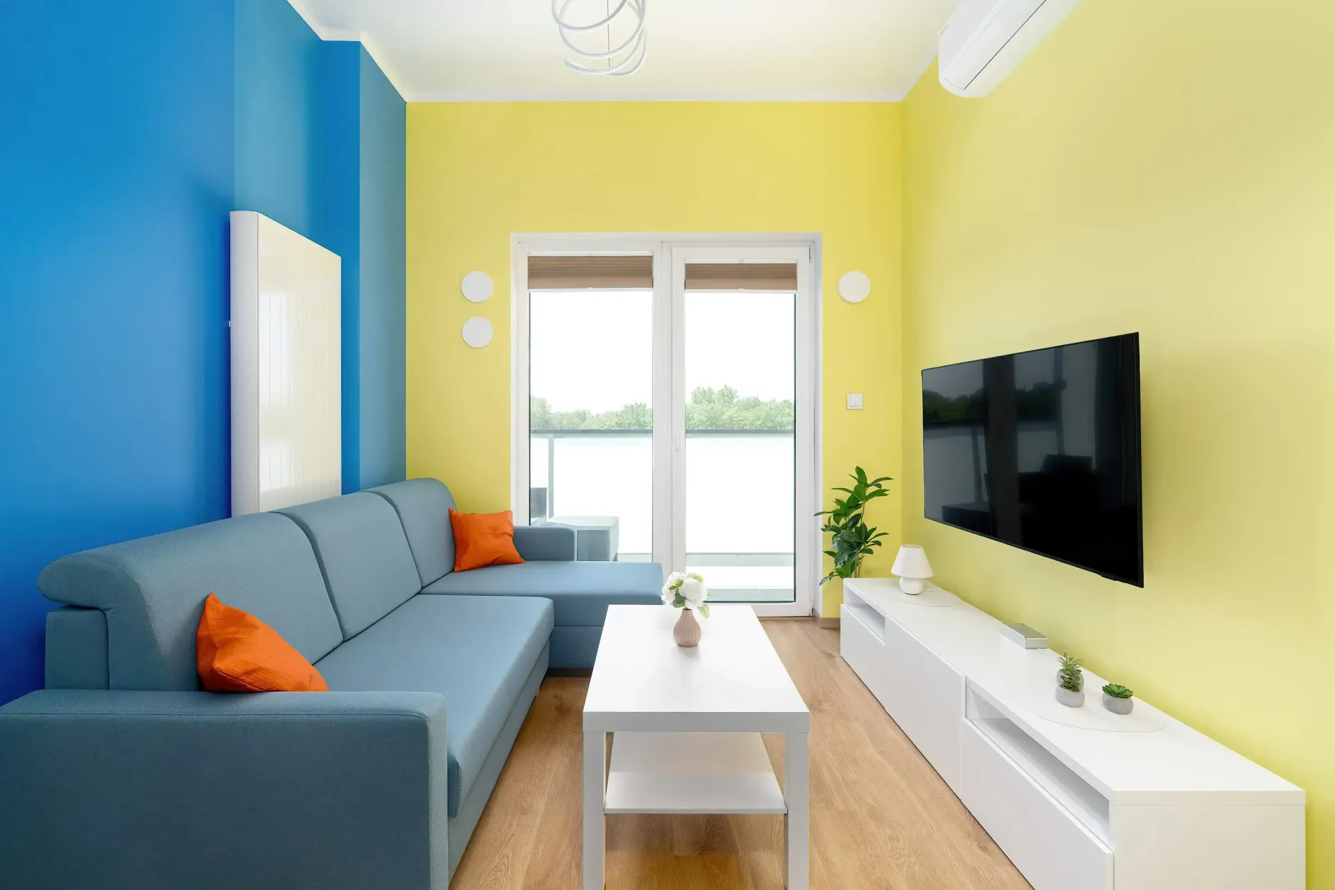Niebieska sofa modułowa w salonie z niebieskimi i żółtymi ścianami, telewizorem i drzwiami balkonowymi.
