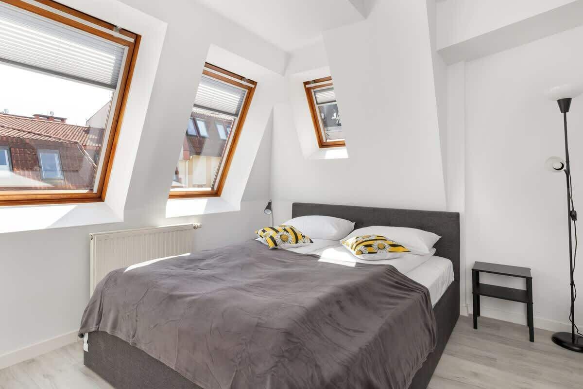 Sypialnia z szarym łóżkiem, białymi ścianami, skośnymi oknami dachowymi, małym czarnym stolikiem nocnym i wysoką lampą podłogową.