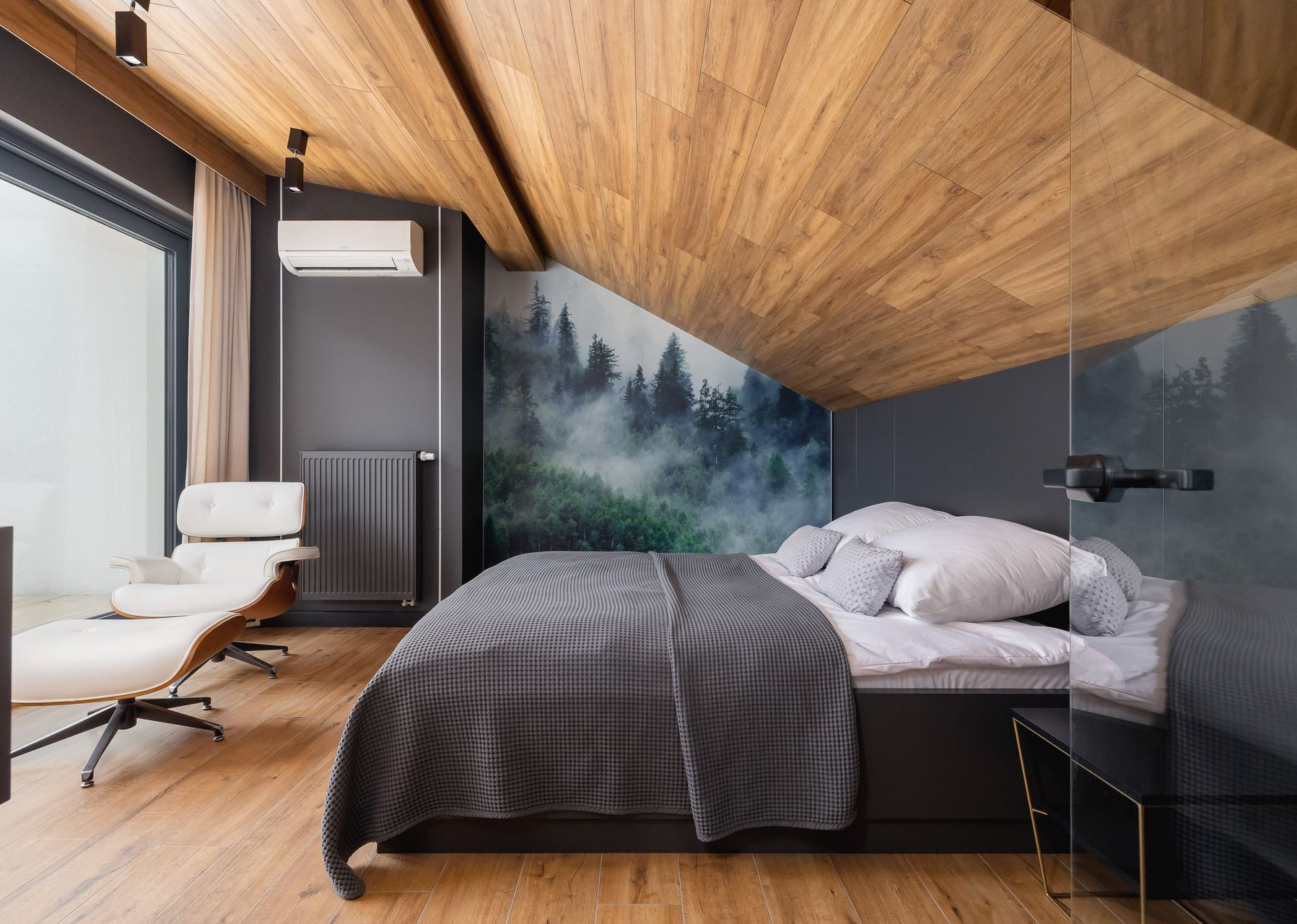 Sypialnia z drewnianym sufitem, łóżkiem z szarym kocem, fototapetą przedstawiającą las, fotelem rozkładanym i dużym oknem.