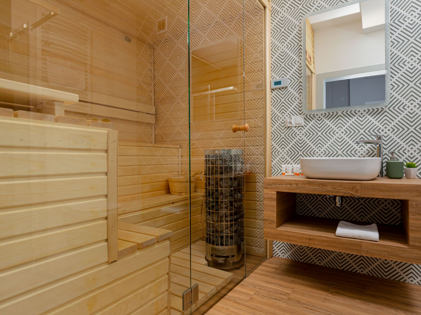 Niewielka sauna ze szklanymi drzwiami, połączona z łazienką wykończoną drewnem i wzorzystymi płytkami.