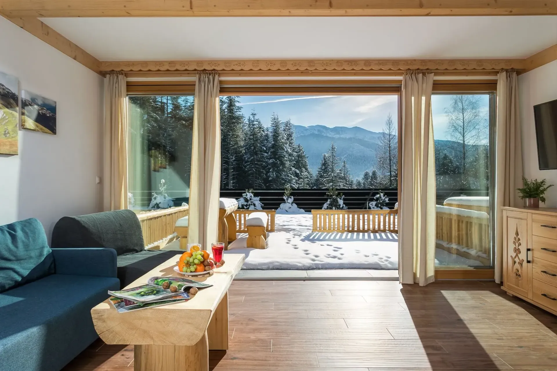 Przytulny salon z otwartymi drzwiami wychodzącymi na ośnieżone góry. Drewniane meble, niebieska sofa i jasna, zimowa sceneria.