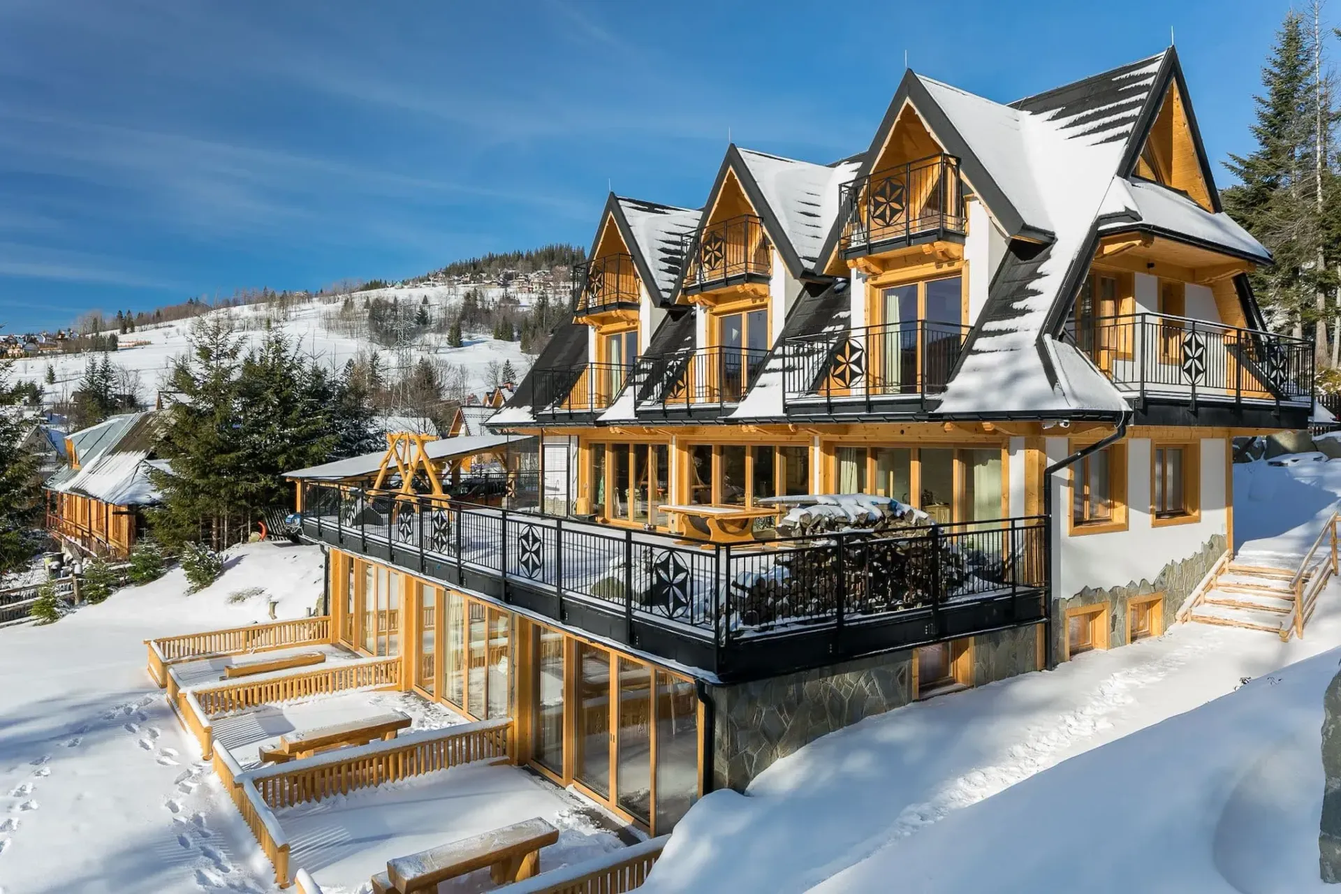 Pokryty śniegiem drewniany domek z balkonami, położony na ośnieżonym zboczu góry, w słoneczny dzień.