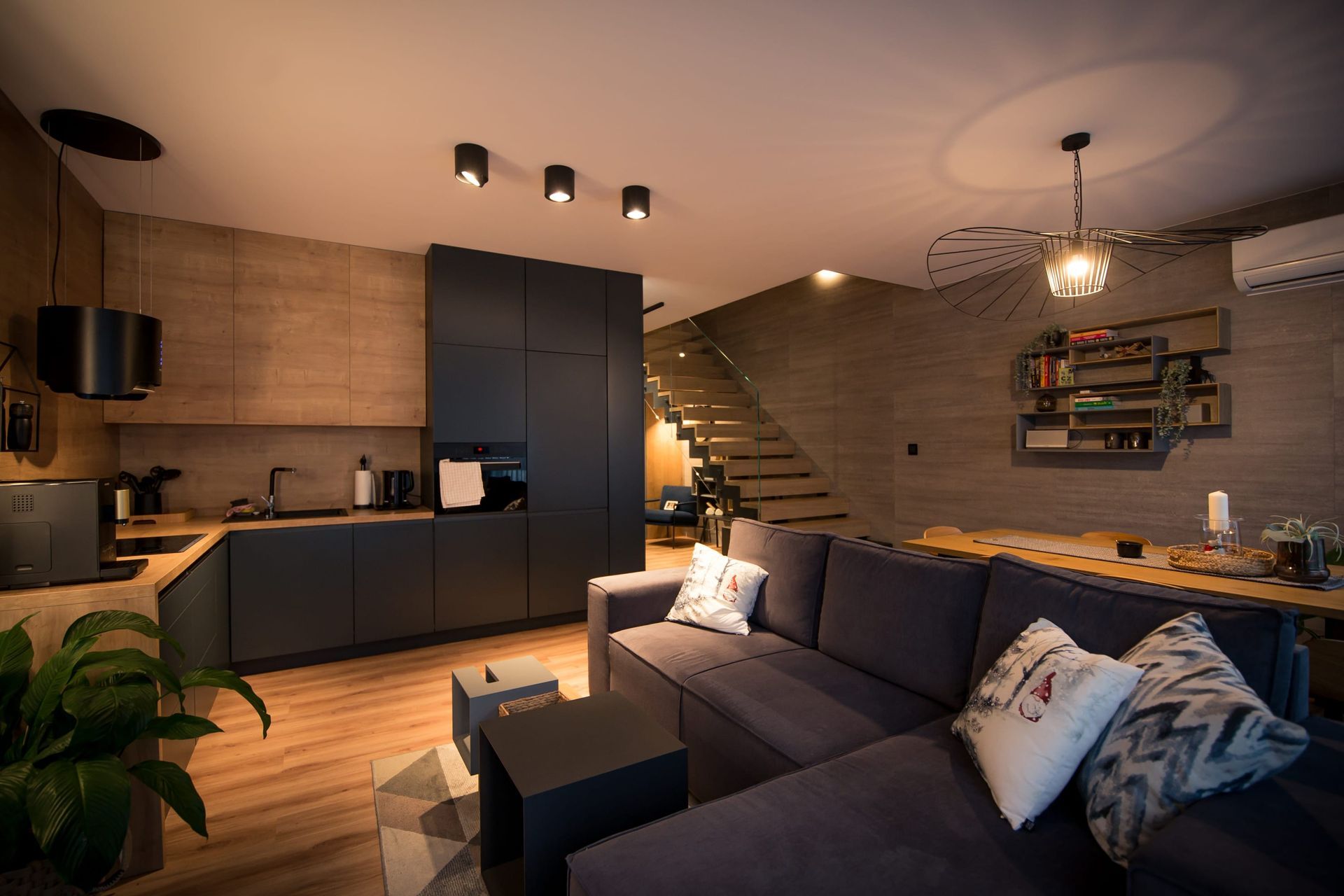Nowoczesny salon z ciemnoszarą narożną sofą, kuchnią i drewnianymi schodami.