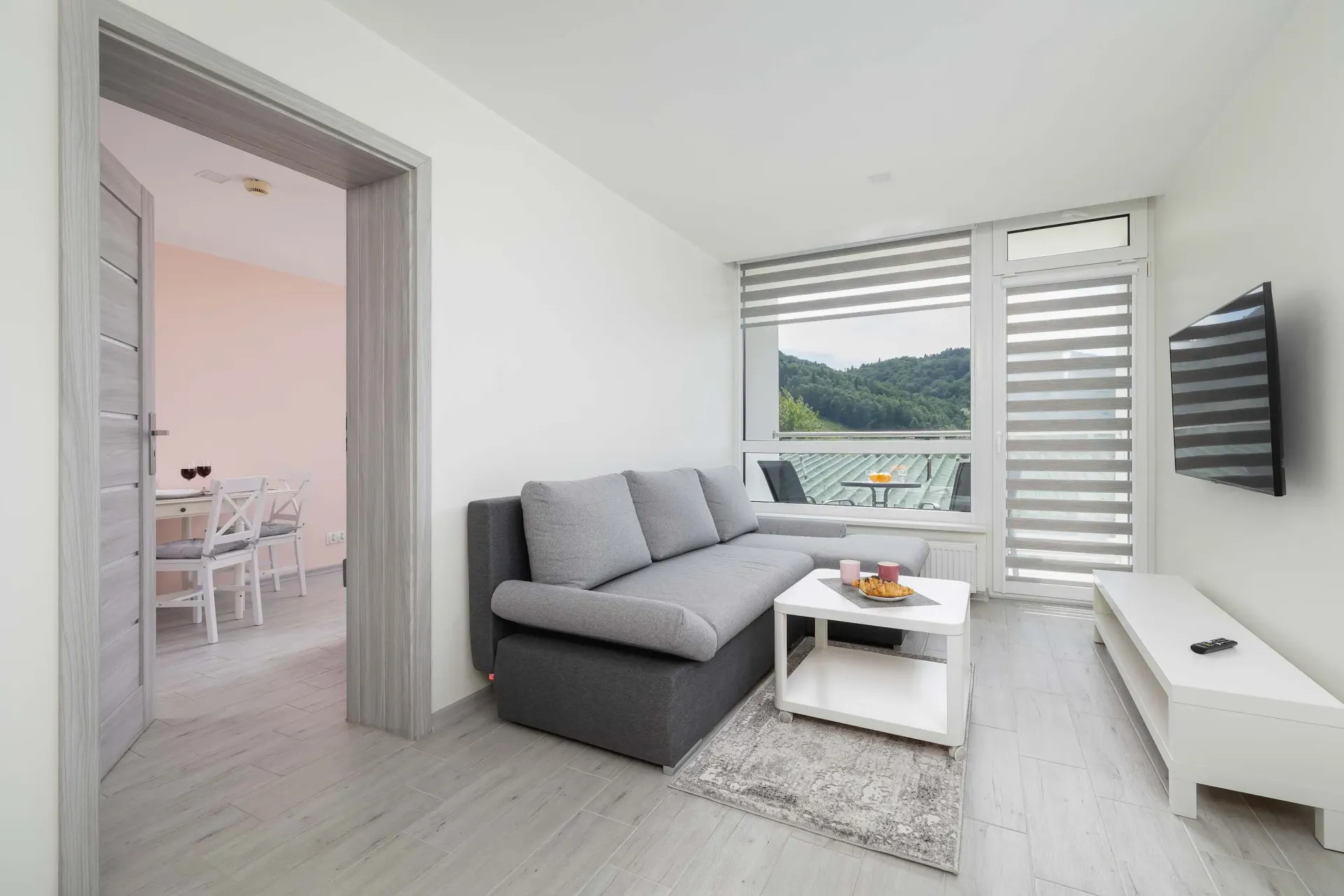 Nowoczesny salon z szarą sofą, białymi ścianami i widokiem przez duże okno.