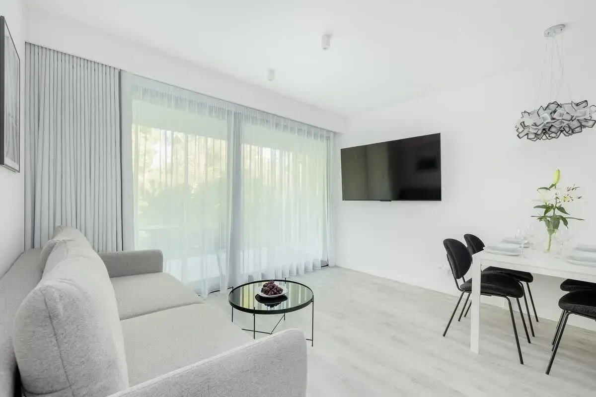 Salon z szarą sofą, cienkimi zasłonami, zamontowanym telewizorem i stołem jadalnym z czarnymi krzesłami.