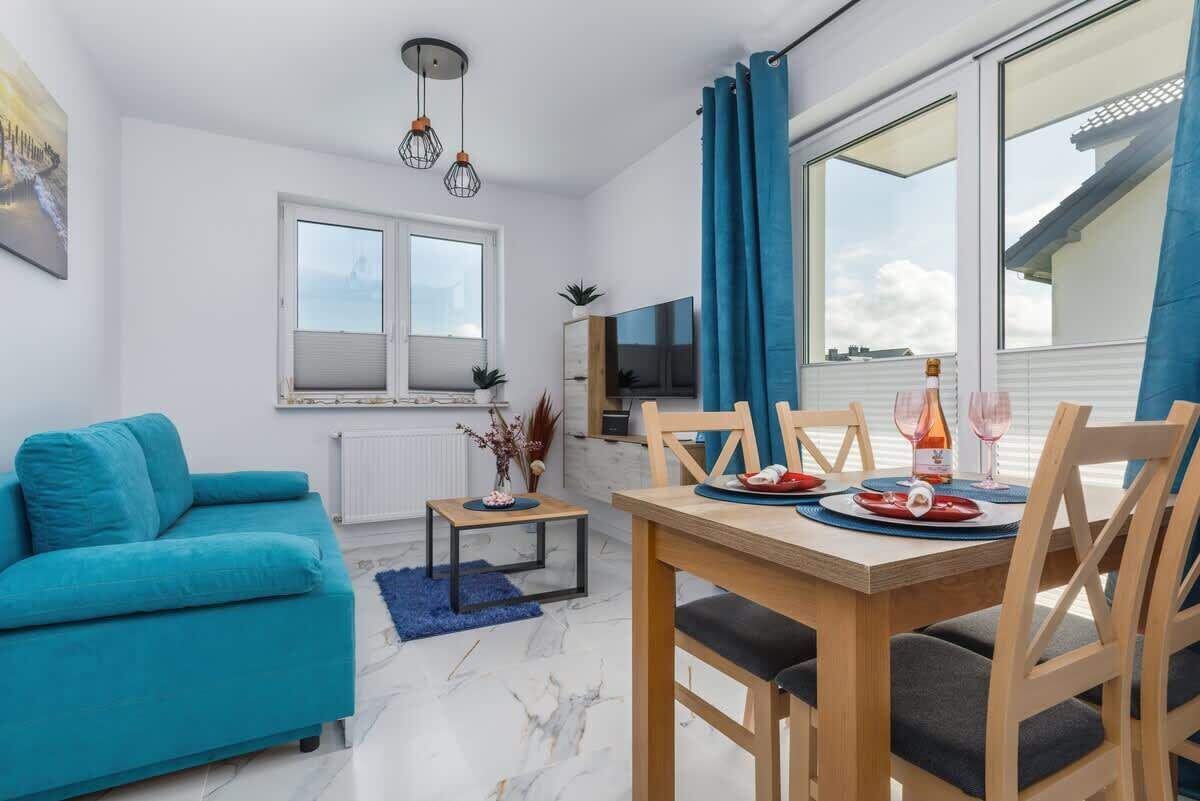 Jasny salon z turkusową sofą, stołem jadalnym i dużym oknem z niebieskimi zasłonami.