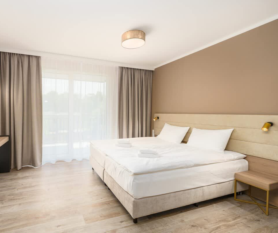 Pokój z starannie pościelonym podwójnym łóżkiem, jasną drewnianą podłogą, telewizorem zamontowanym na ścianie i beżowymi zasłonami sięgającymi od podłogi do sufitu.