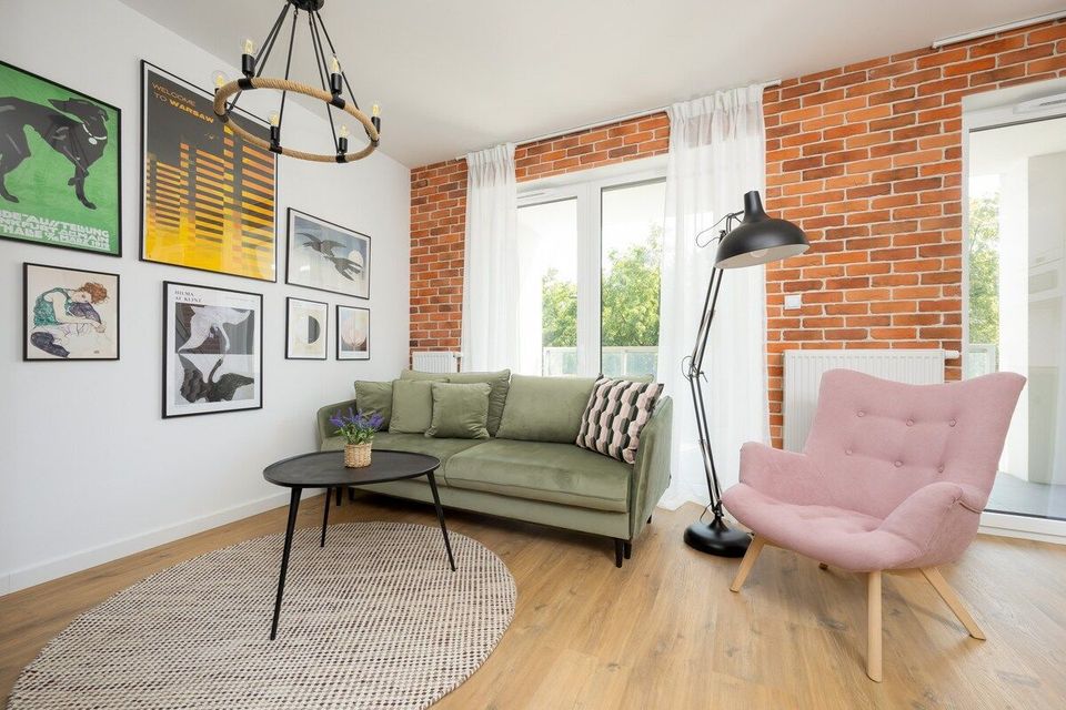 Jasny salon z zieloną sofą, różowym fotelem, stolikiem kawowym, ceglanymi ścianami i dużymi oknami