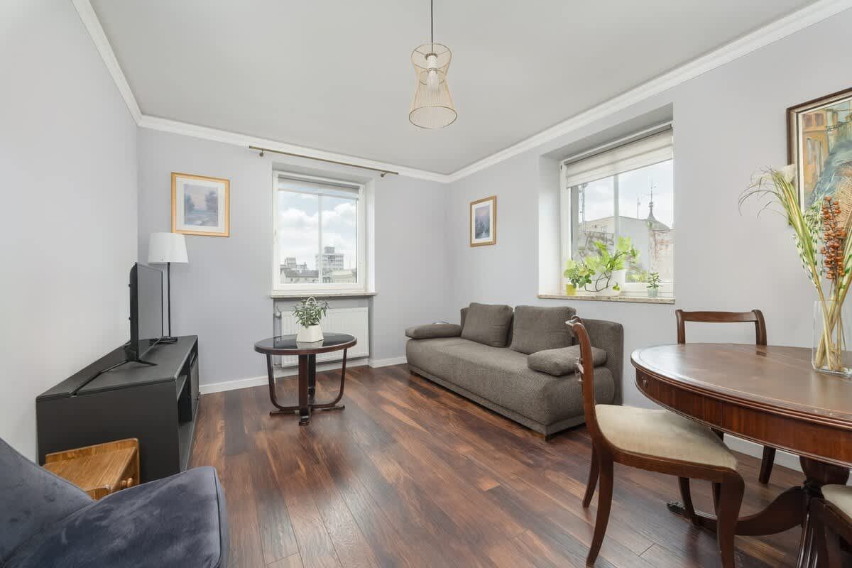 Salon z drewnianą podłogą, szarymi ścianami, sofą, stołem i dwoma oknami.