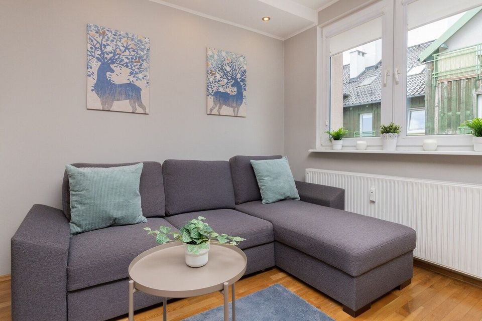 Szara sofa modułowa w jasnym salonie z niebieskimi poduszkami, grafiką na ścianie i małym okrągłym stolikiem kawowym.