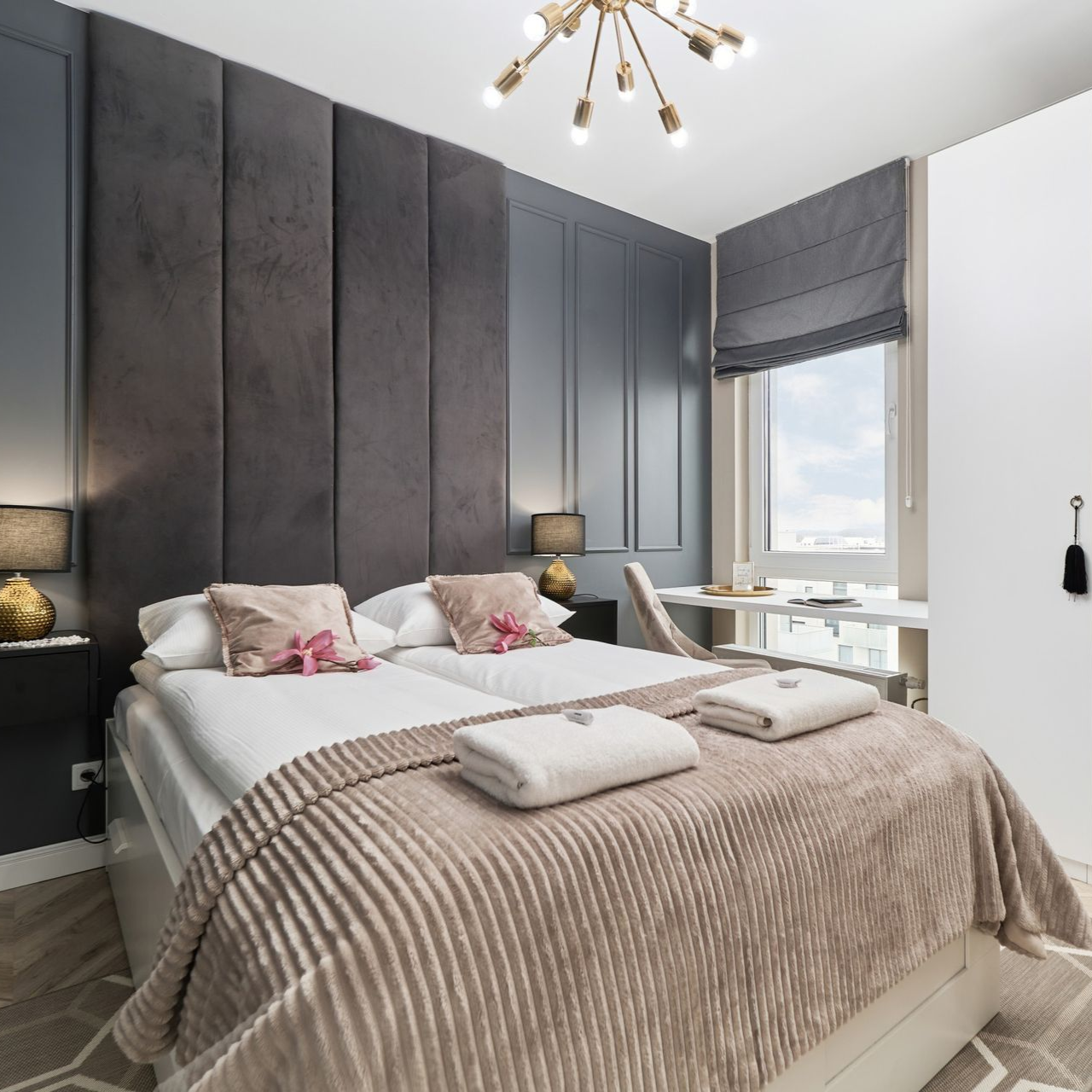 Schlafzimmer mit Doppelbett, grauer Wandvertäfelung und einem kleinen Schreibtisch am Fenster mit einem römischen Rollo.